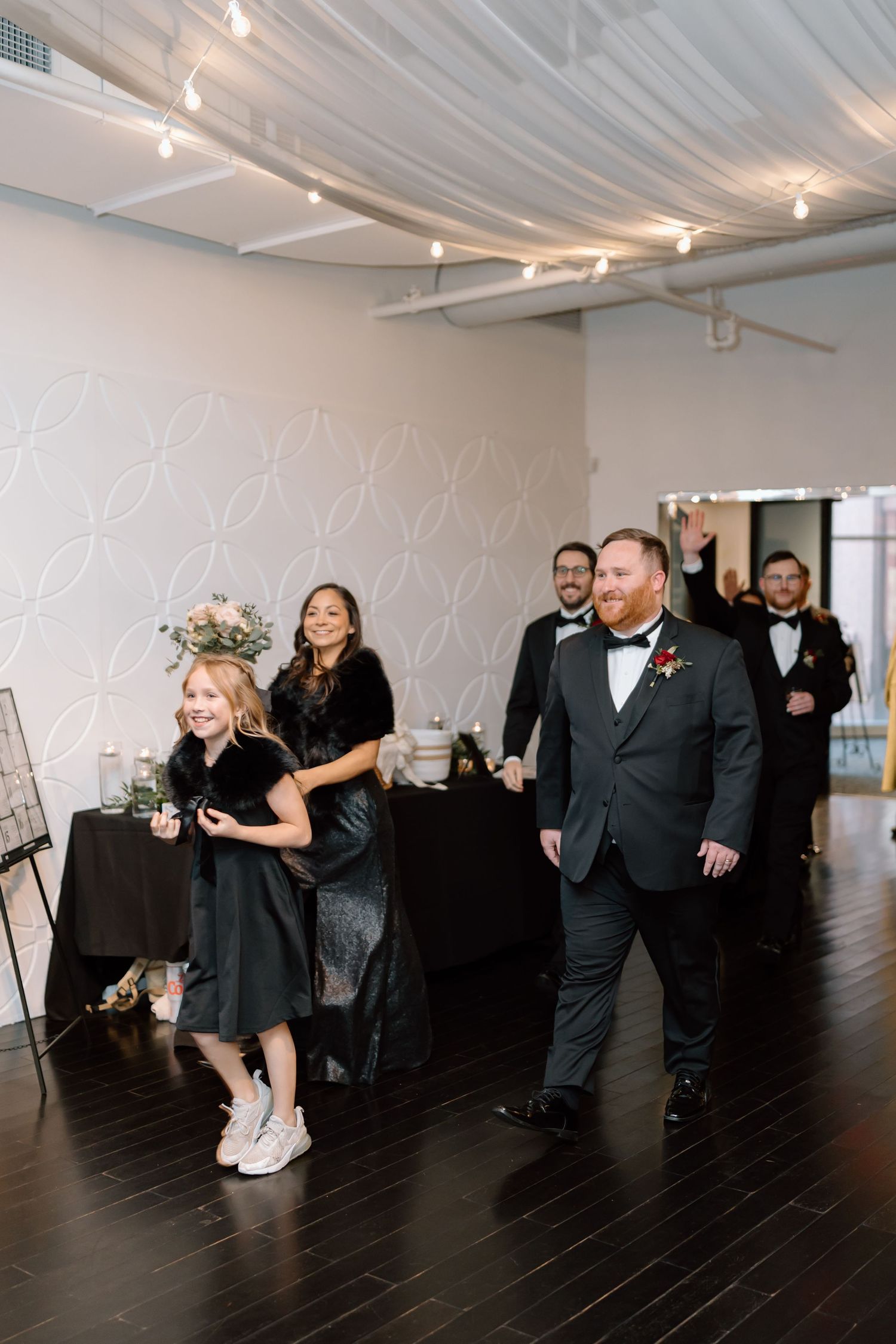 Wedding guests dance together on a dark hardwood floor in a white-walled venue with track lighting.