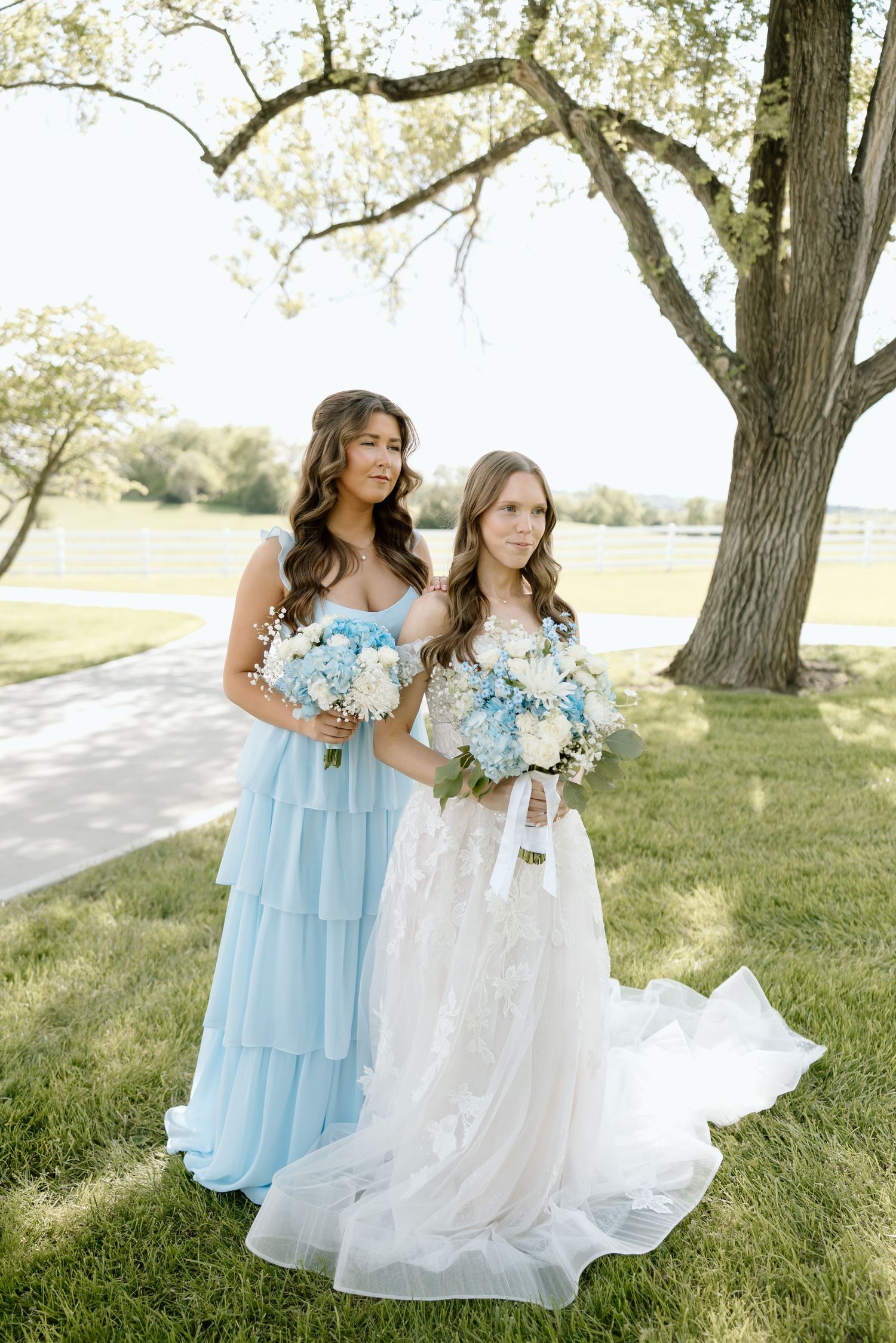 Bride in lace gown and bridesmaid in tiered blue dress pose with matching bouquets under large tree.
