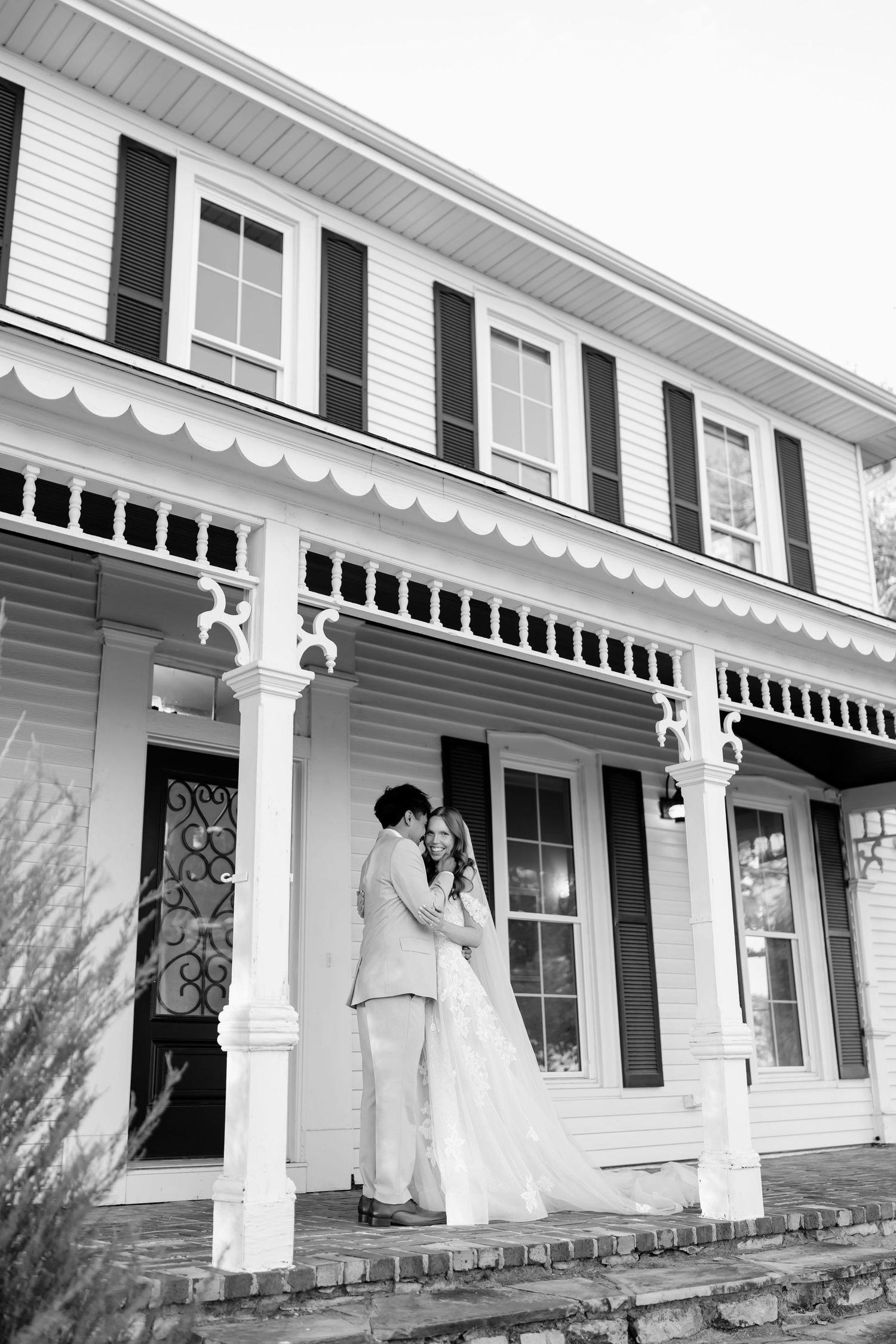 Black and white photo of a couple embracing on the porch of a classic white colonial home during their wedding day.