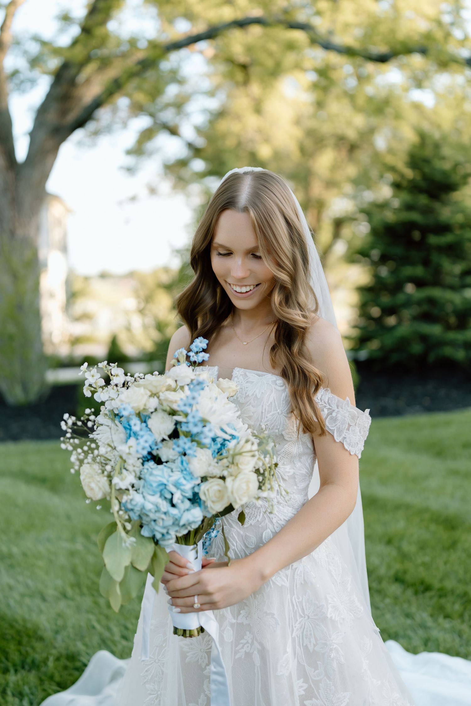 Smiling bride in lace off-shoulder gown holds blue and white floral bouquet outdoors on green lawn.