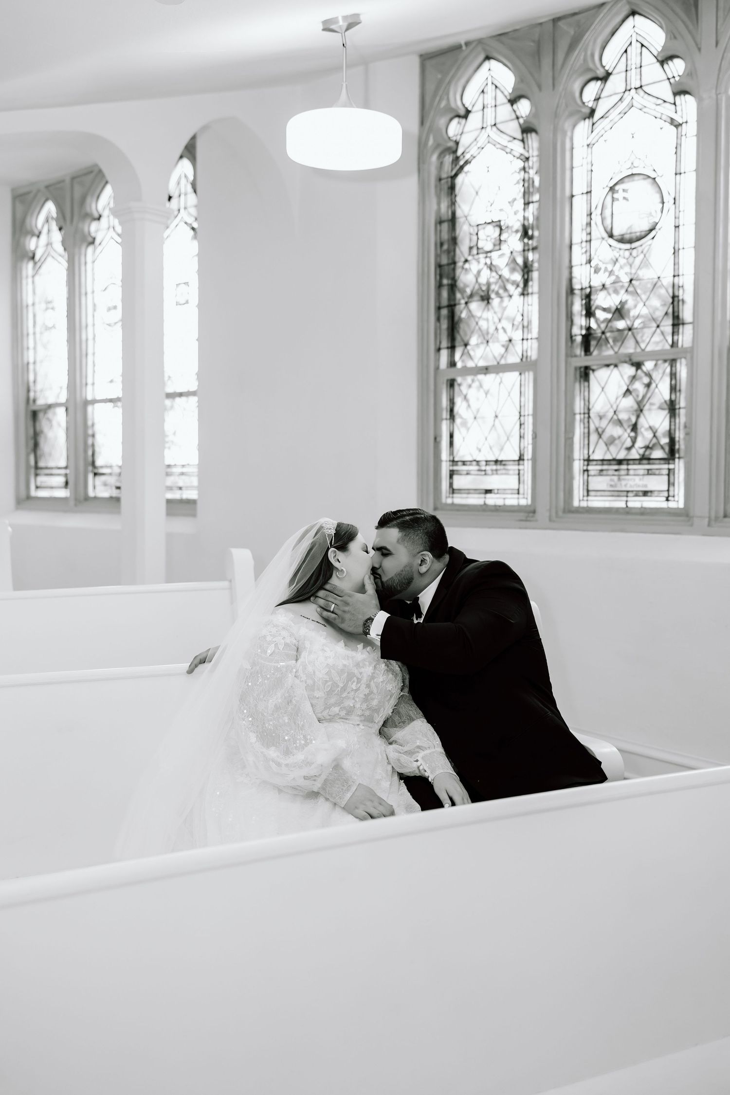 Black and white photo of a bride and groom sharing a kiss in a church pew with stained glass windows behind them.
