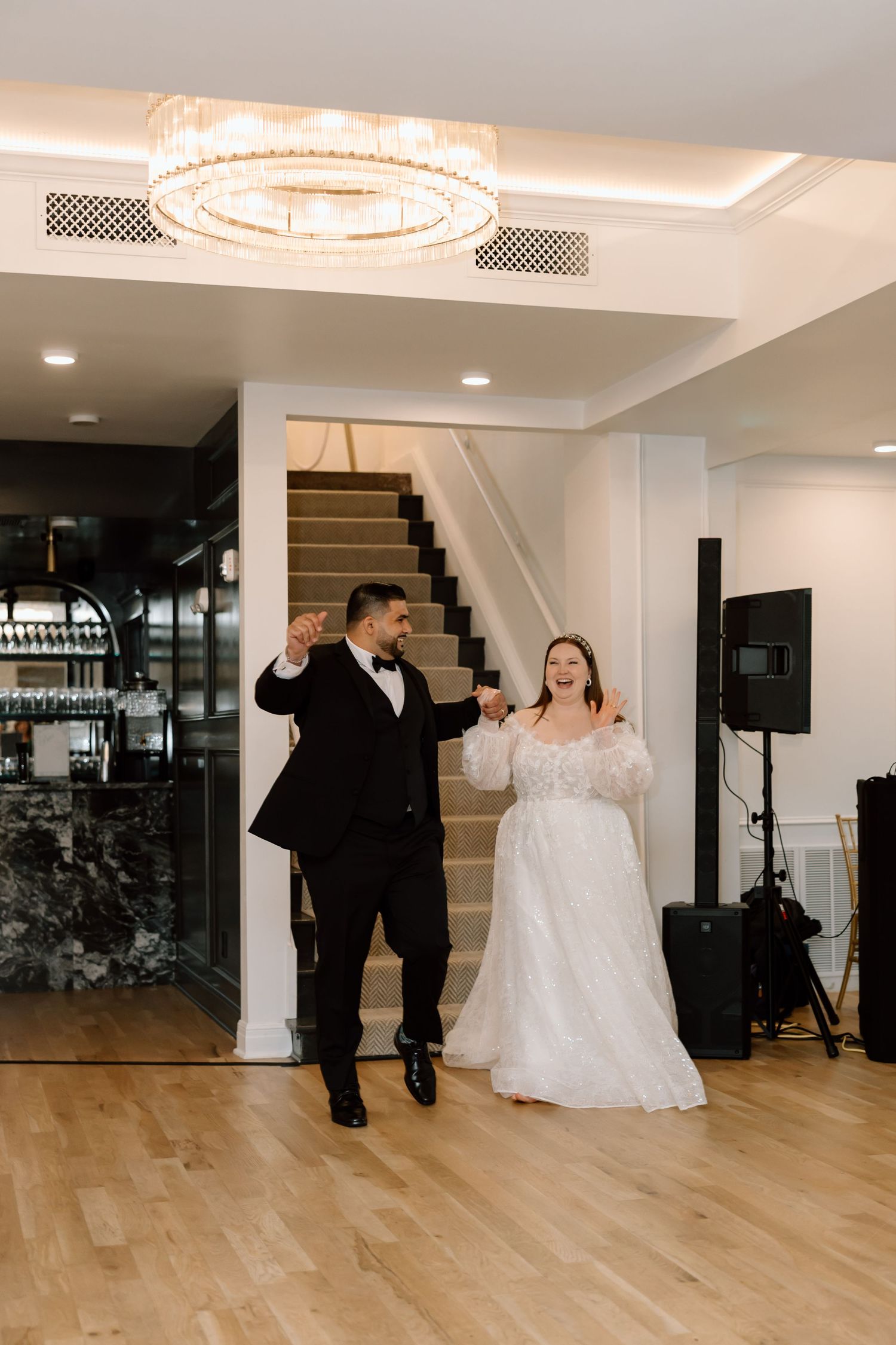 Bride and groom make grand entrance at wedding reception, dancing joyfully under elegant chandelier on hardwood floor.