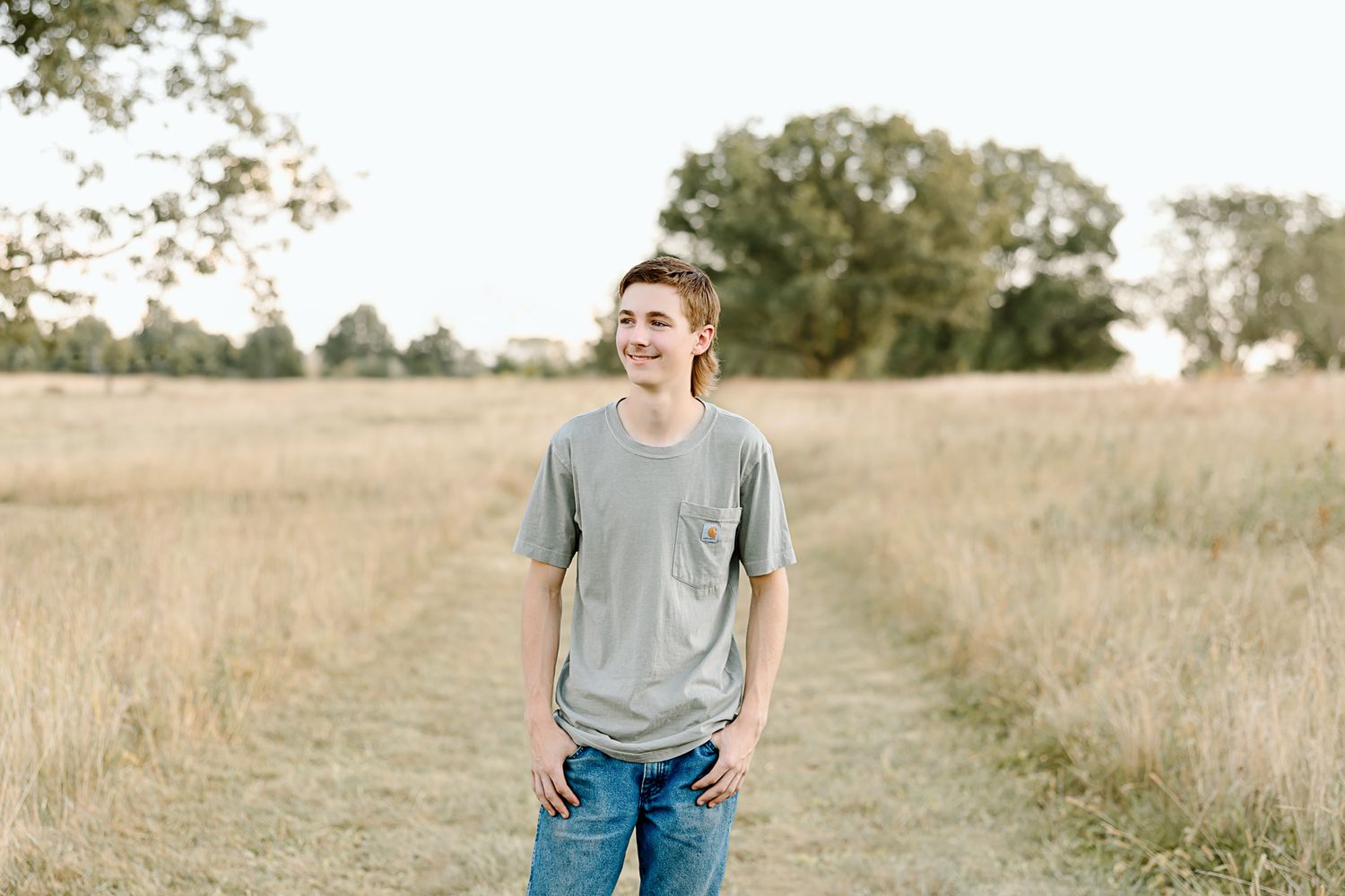 Young person in gray t-shirt and jeans standing in golden field at sunset.