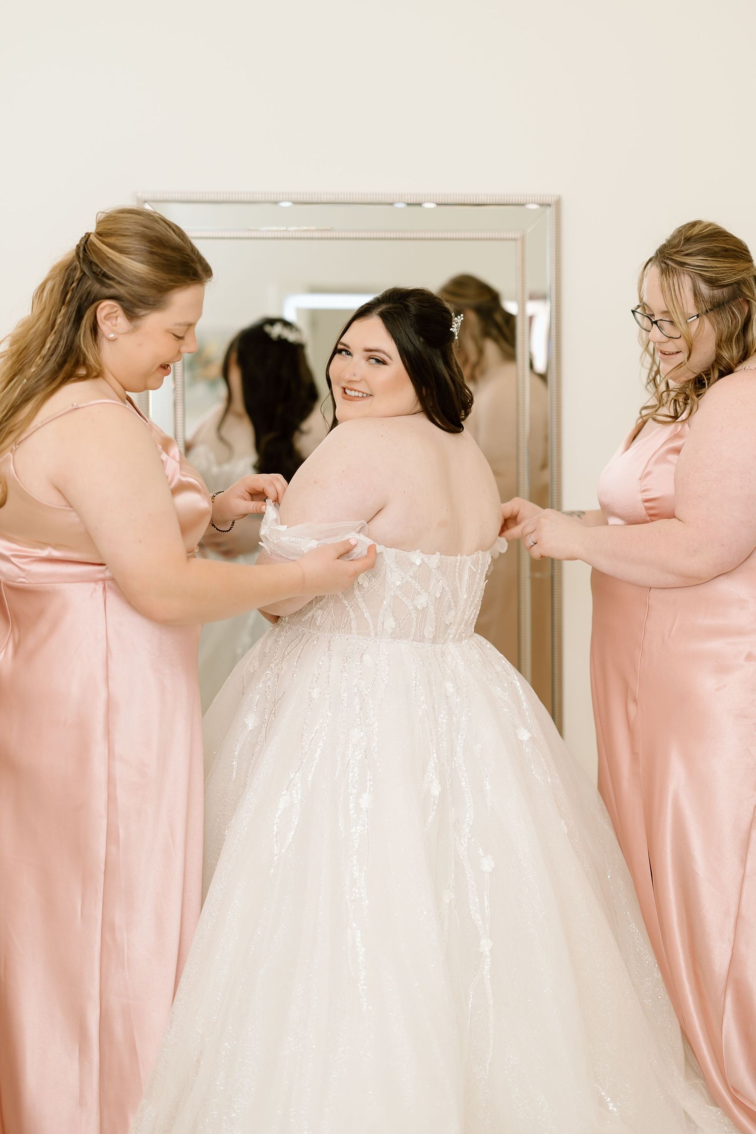 Bridesmaids in pink dresses help bride with wedding gown preparations.