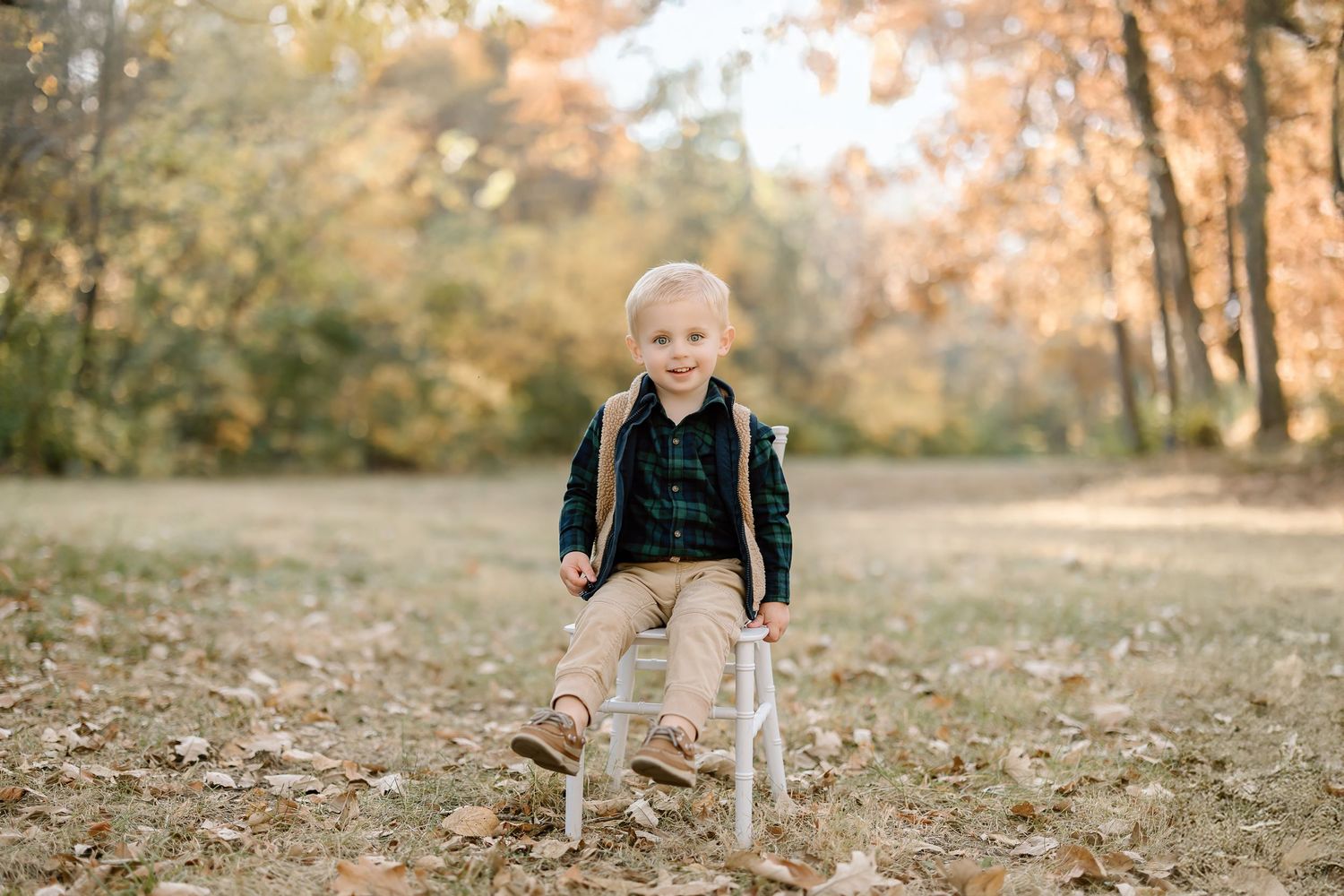 Child in plaid shirt and khaki pants sits on white chair in autumn setting.