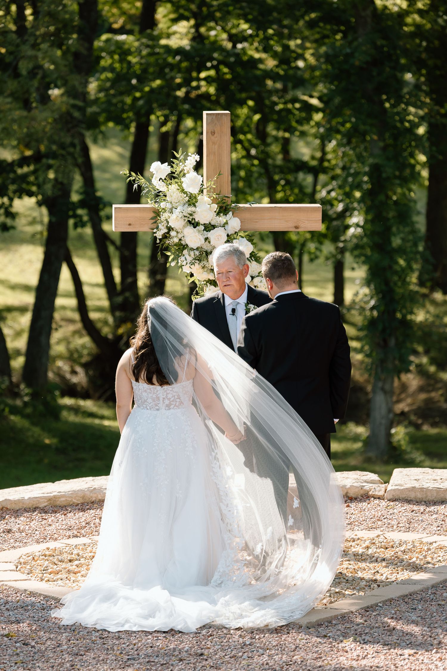 A couple in wedding attire walks along a sunlit wooded path during their outdoor ceremony.