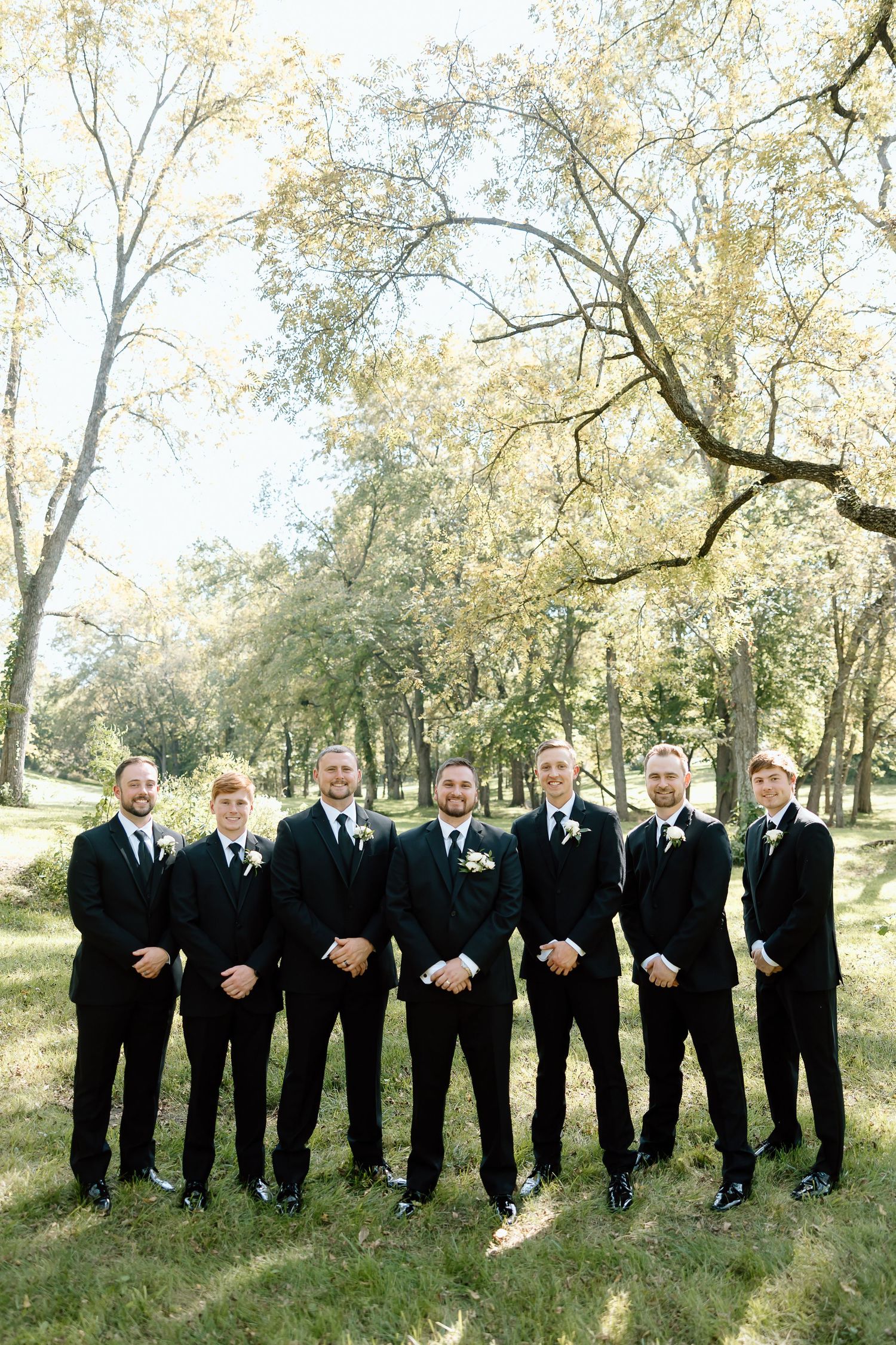 Wedding party wearing black suits stands in line outdoors with trees and sunlight in background.