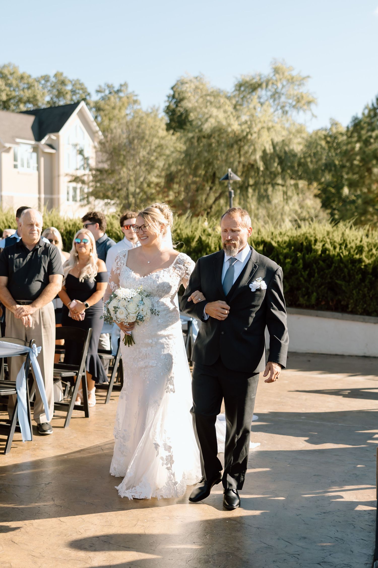 A wedding procession walking down an outdoor patio with guests looking on during a sunny afternoon ceremony.