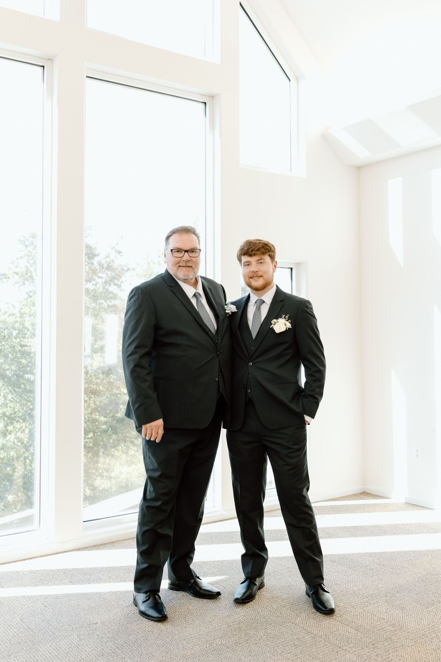 Two people in black suits stand together in a bright room with large windows on a special occasion.