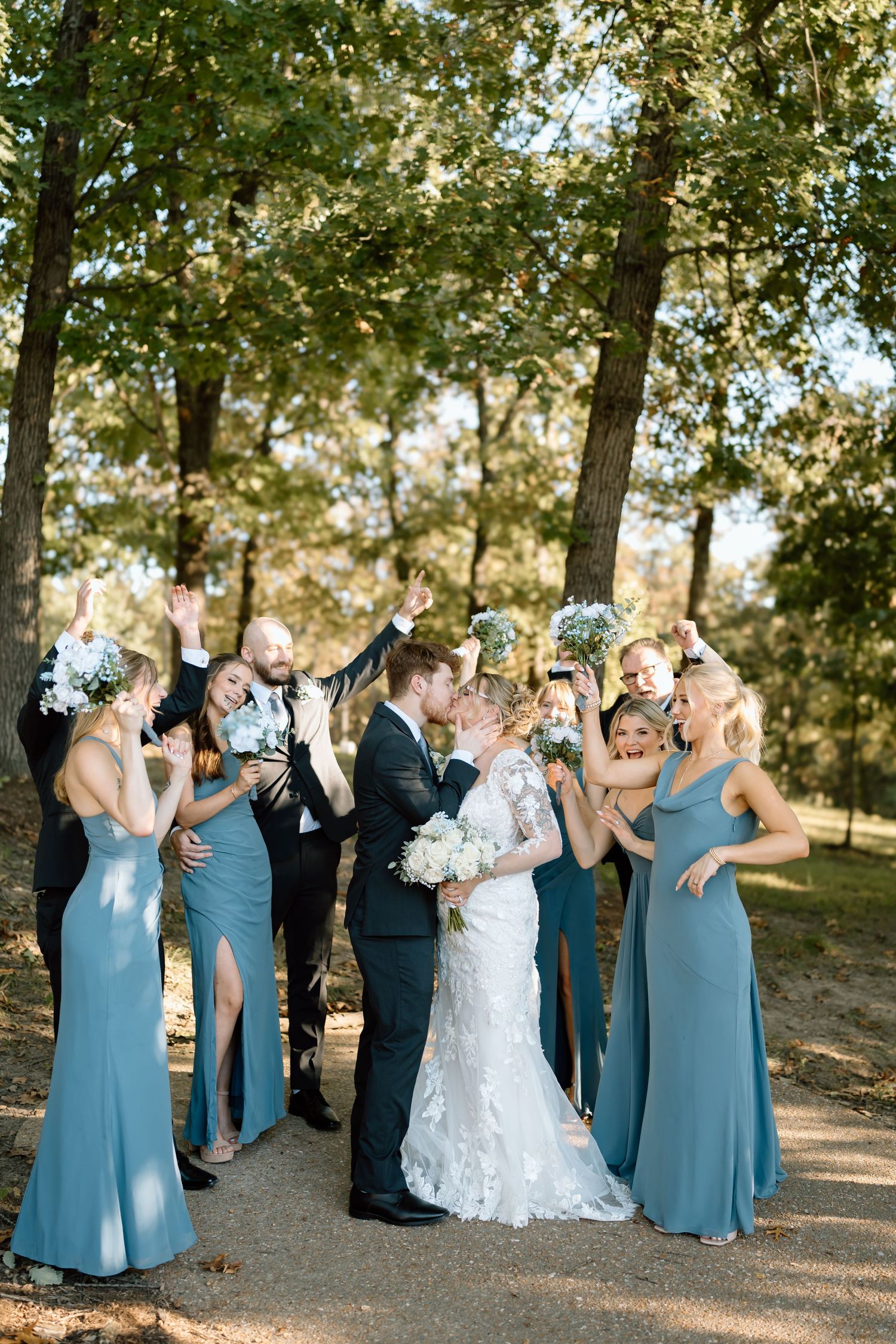 Wedding party celebrates outdoors with bridesmaids in dusty blue dresses and groomsmen in black suits on a sunny day.