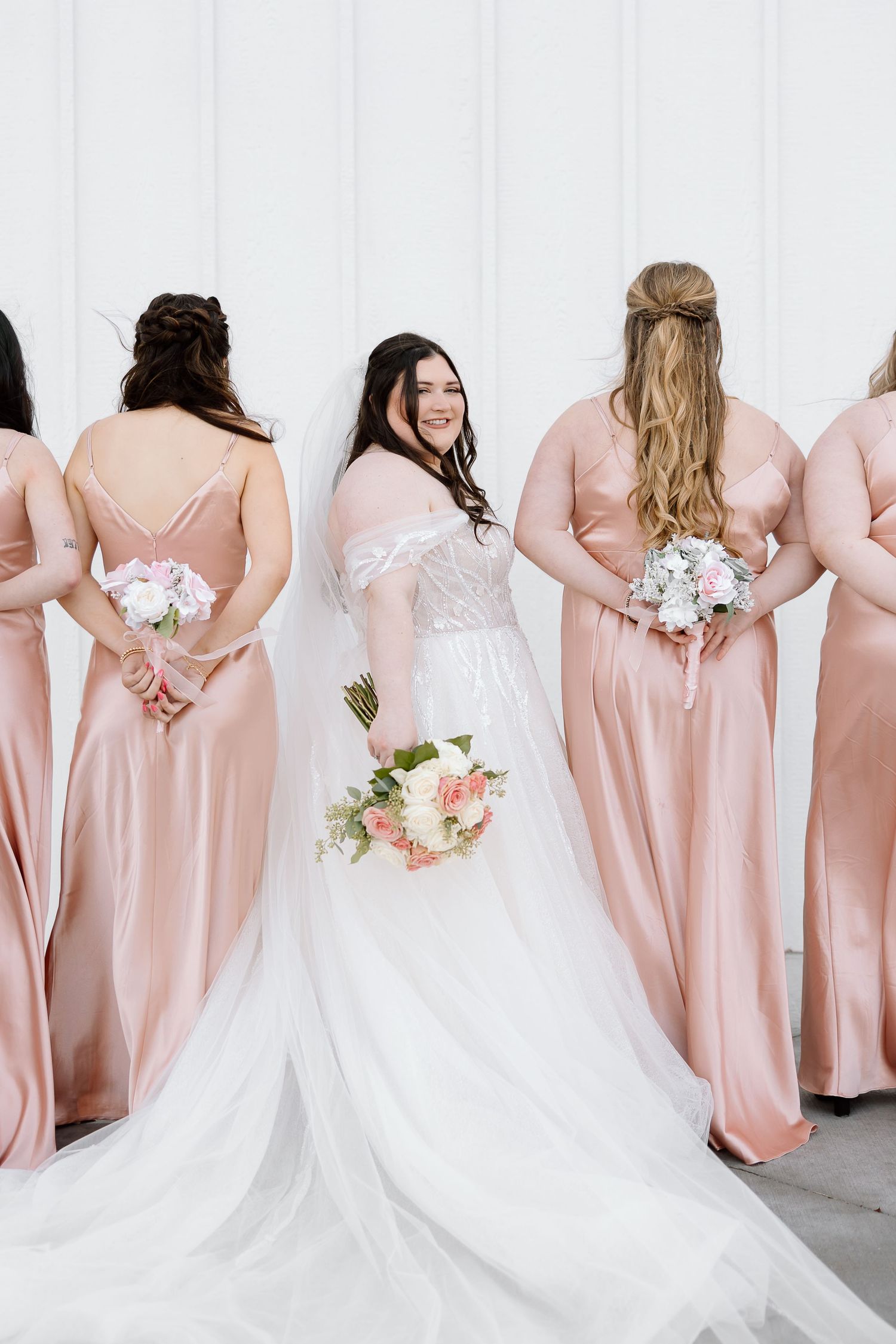 Bridesmaids in blush pink dresses and bride in white gown pose with their backs to camera holding floral bouquets.