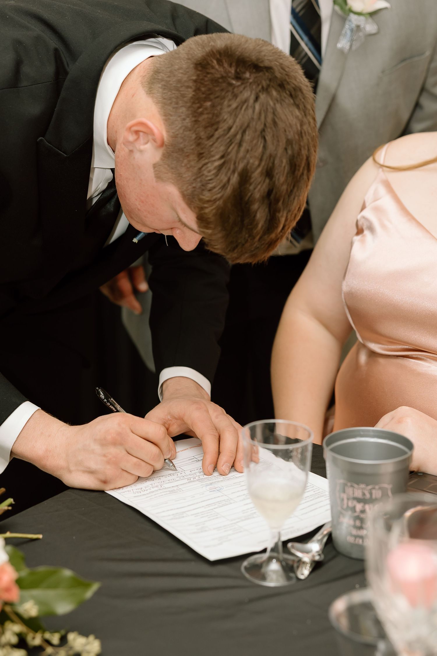 A person in a black suit signs a document at a formal event table with glassware and decorations.