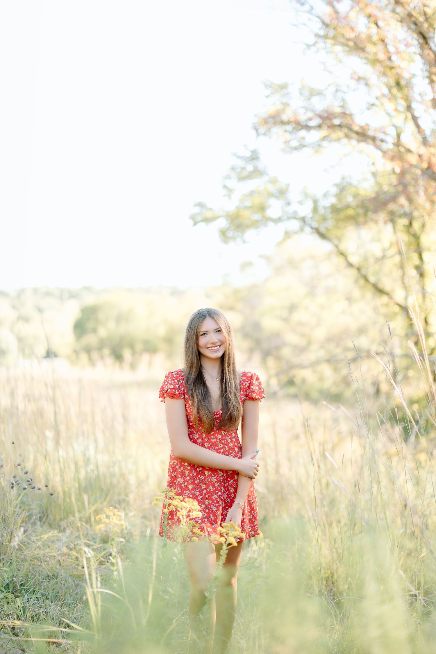 A person in a red floral dress sits gracefully among tall grass in a sunlit meadow during golden hour.