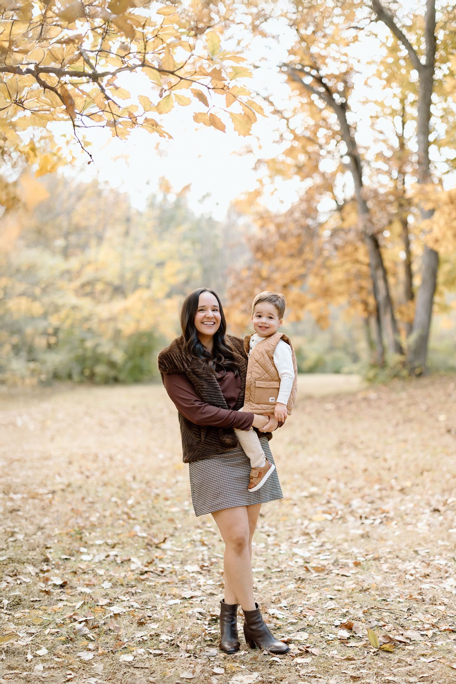 Mother and child playing together in autumn park with fallen golden leaves on ground during outdoor photo session.