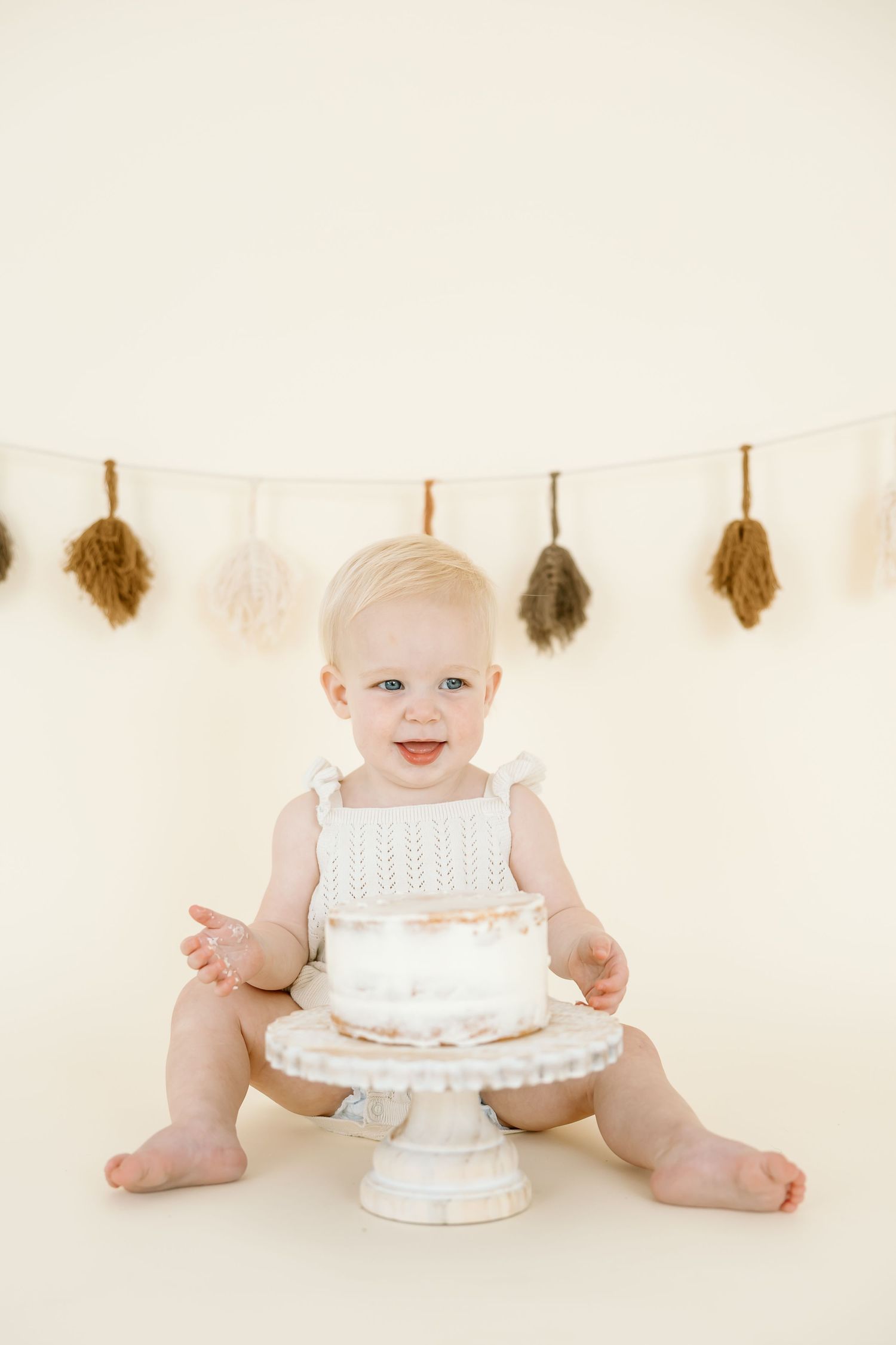 A baby in white sits on a cream backdrop with a small decorated cake on a pedestal stand and feather garland above.