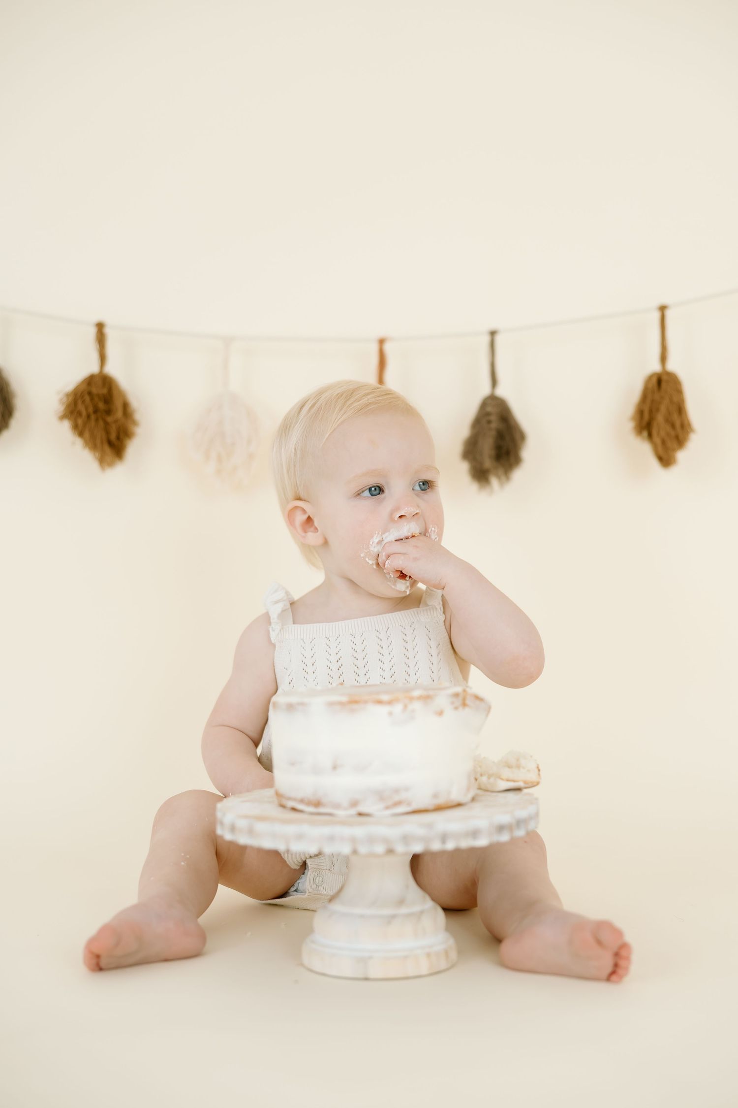 Baby in white sitting on floor with white cake smash setup and neutral feather garland backdrop.