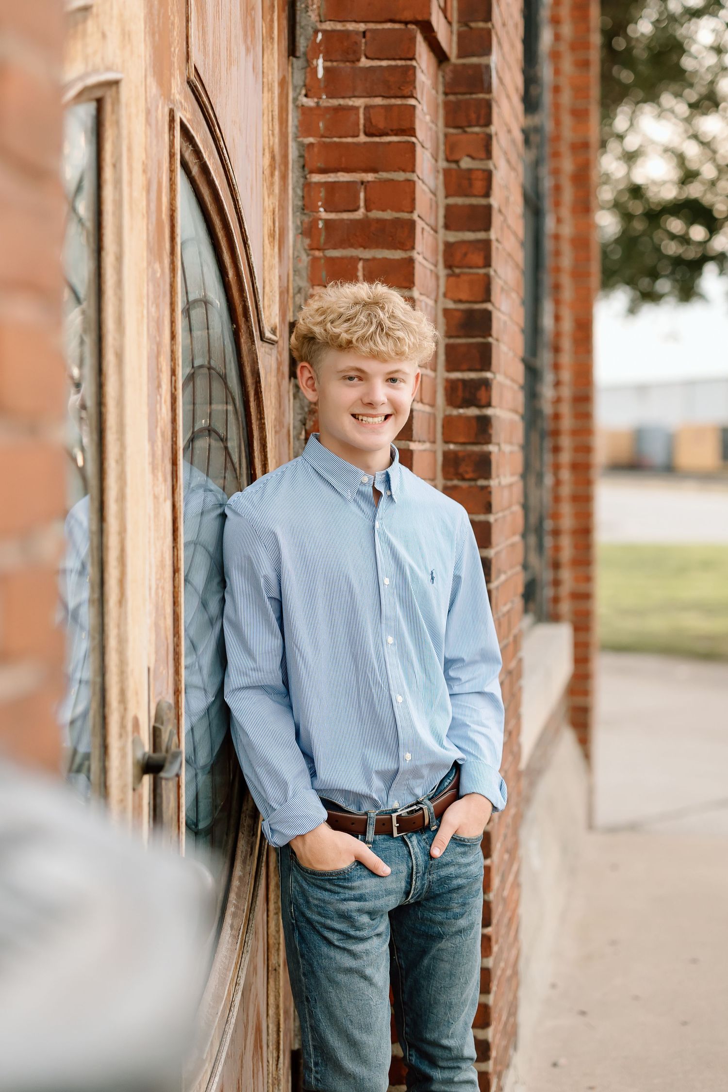 A young boy in a light blue striped shirt and jeans leans against a brick wall in a casual outdoor portrait series.