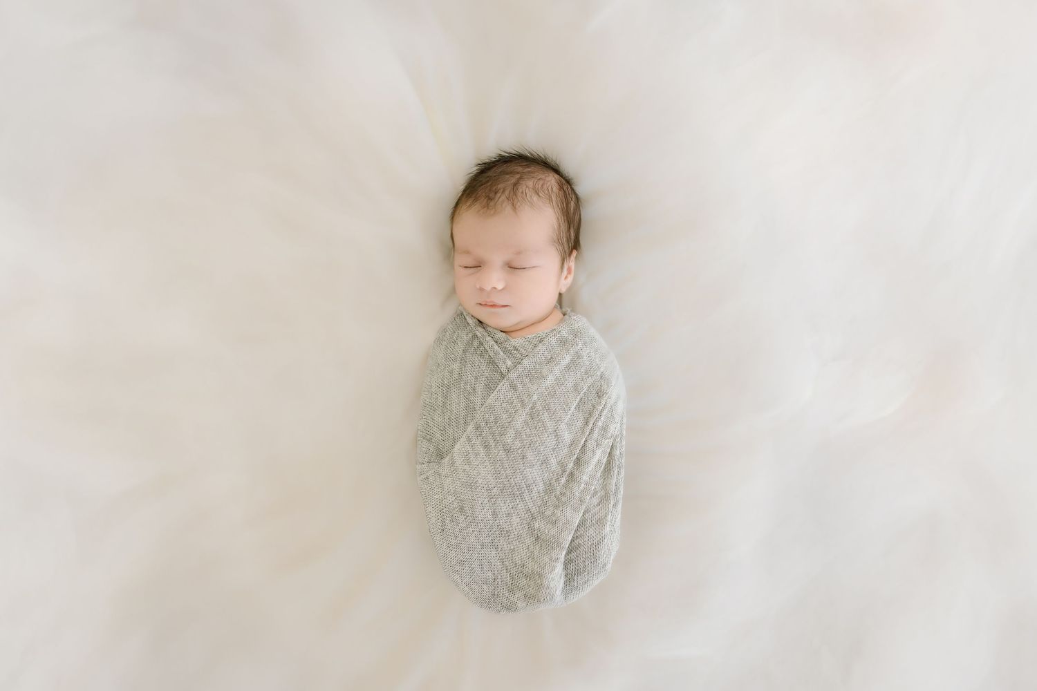 A sleeping newborn baby wrapped in a soft gray swaddle blanket lies peacefully against a white backdrop.