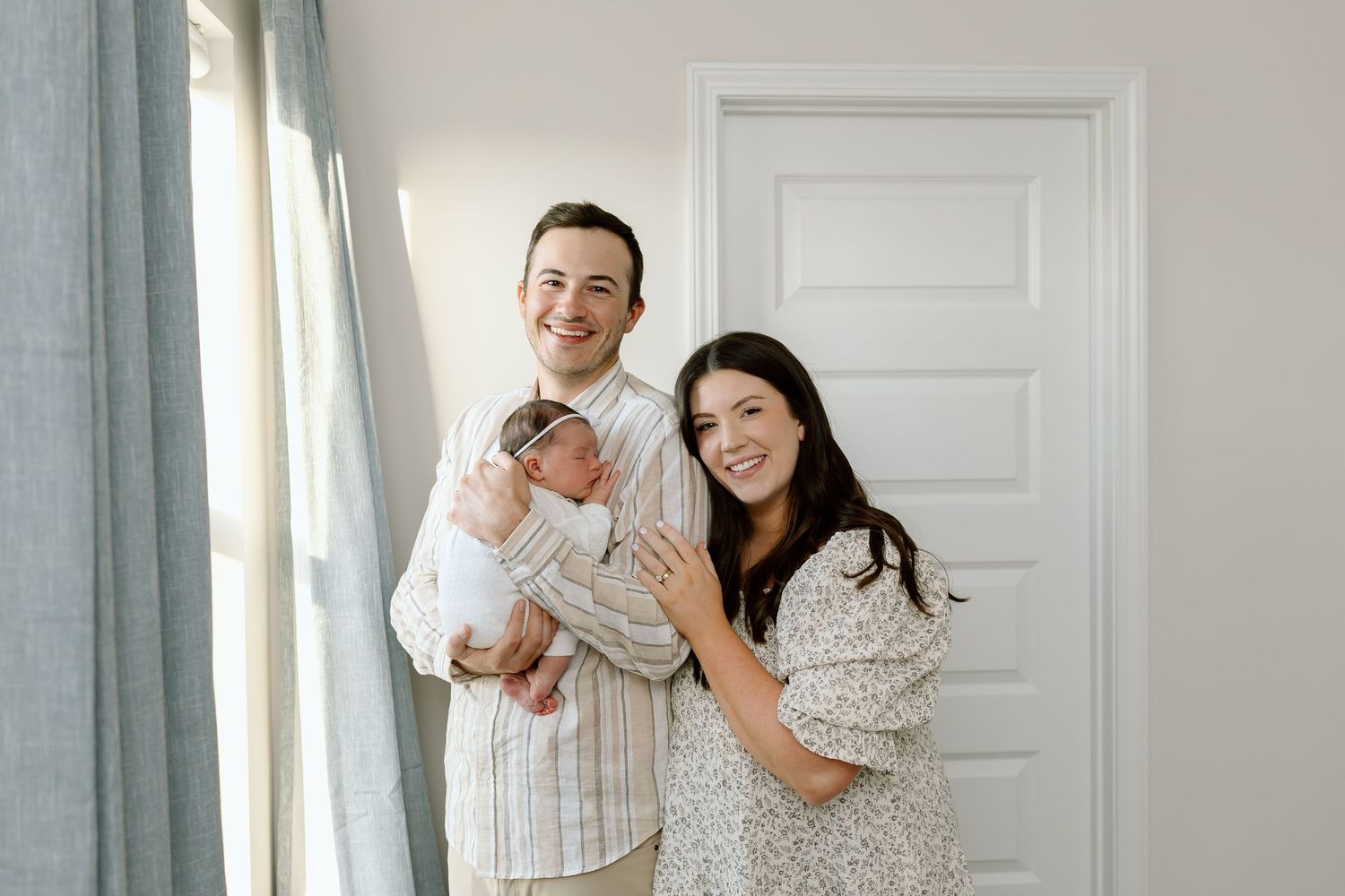 A family poses together in a bright room with white walls and gray curtains.