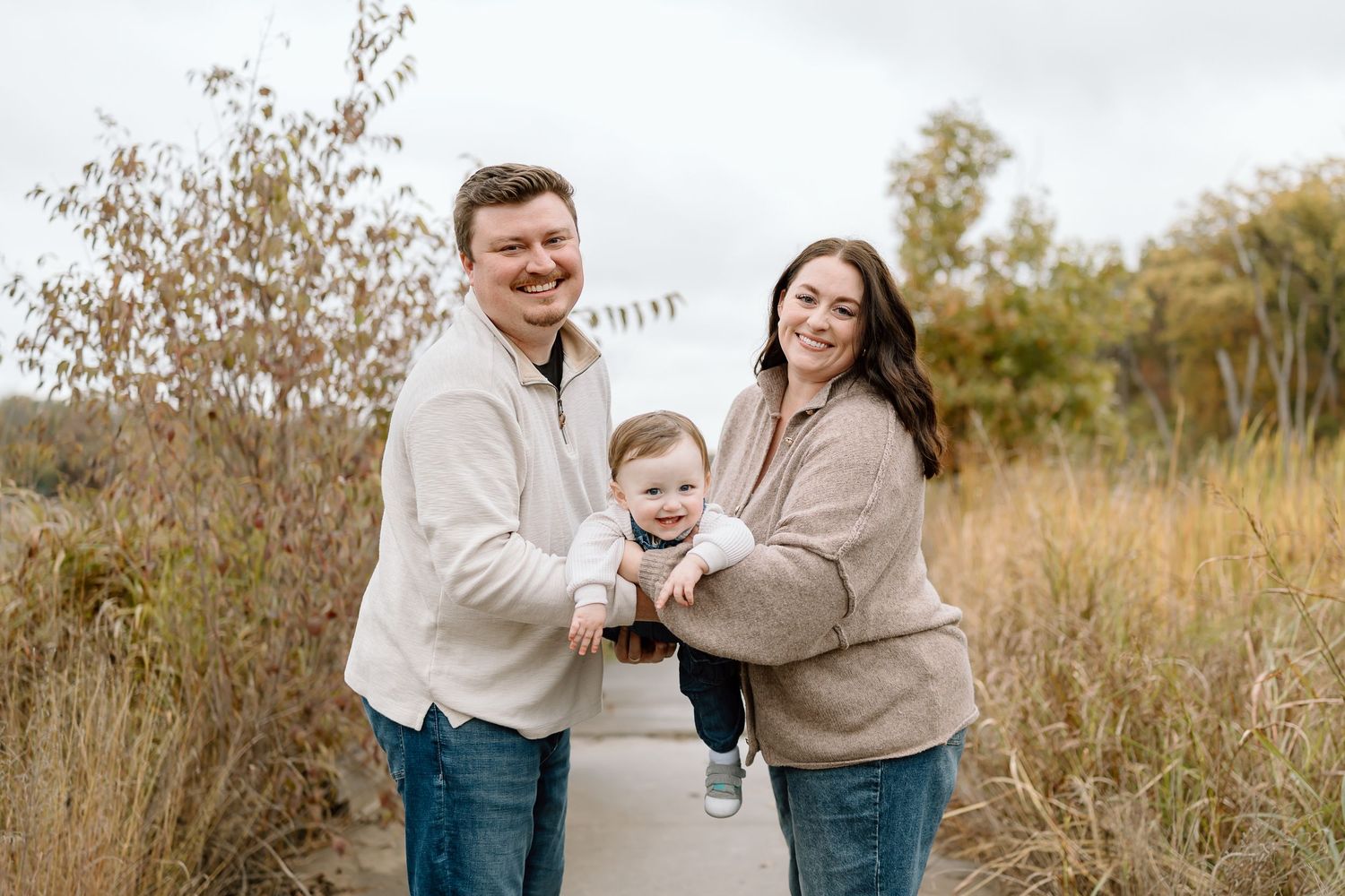 Family wearing coordinated cream colored outfits embraces while laughing together in an autumn field at sunset.