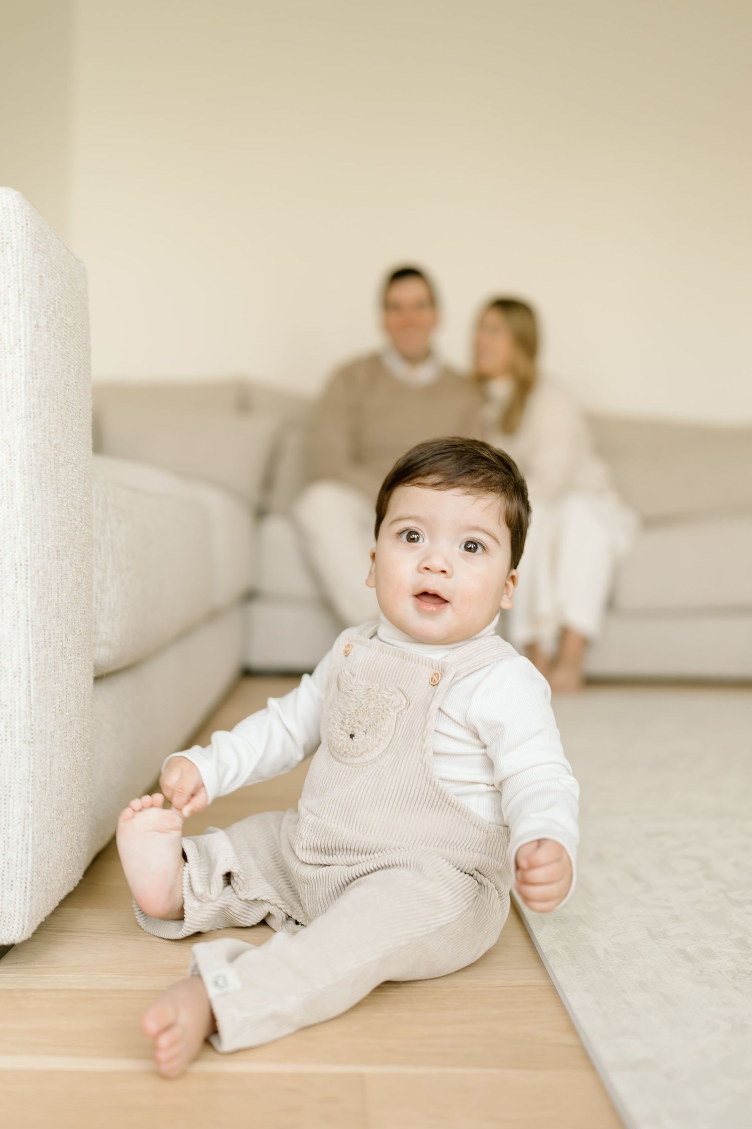 A baby in light beige overalls sits on a hardwood floor with a sofa and two people visible in the background.