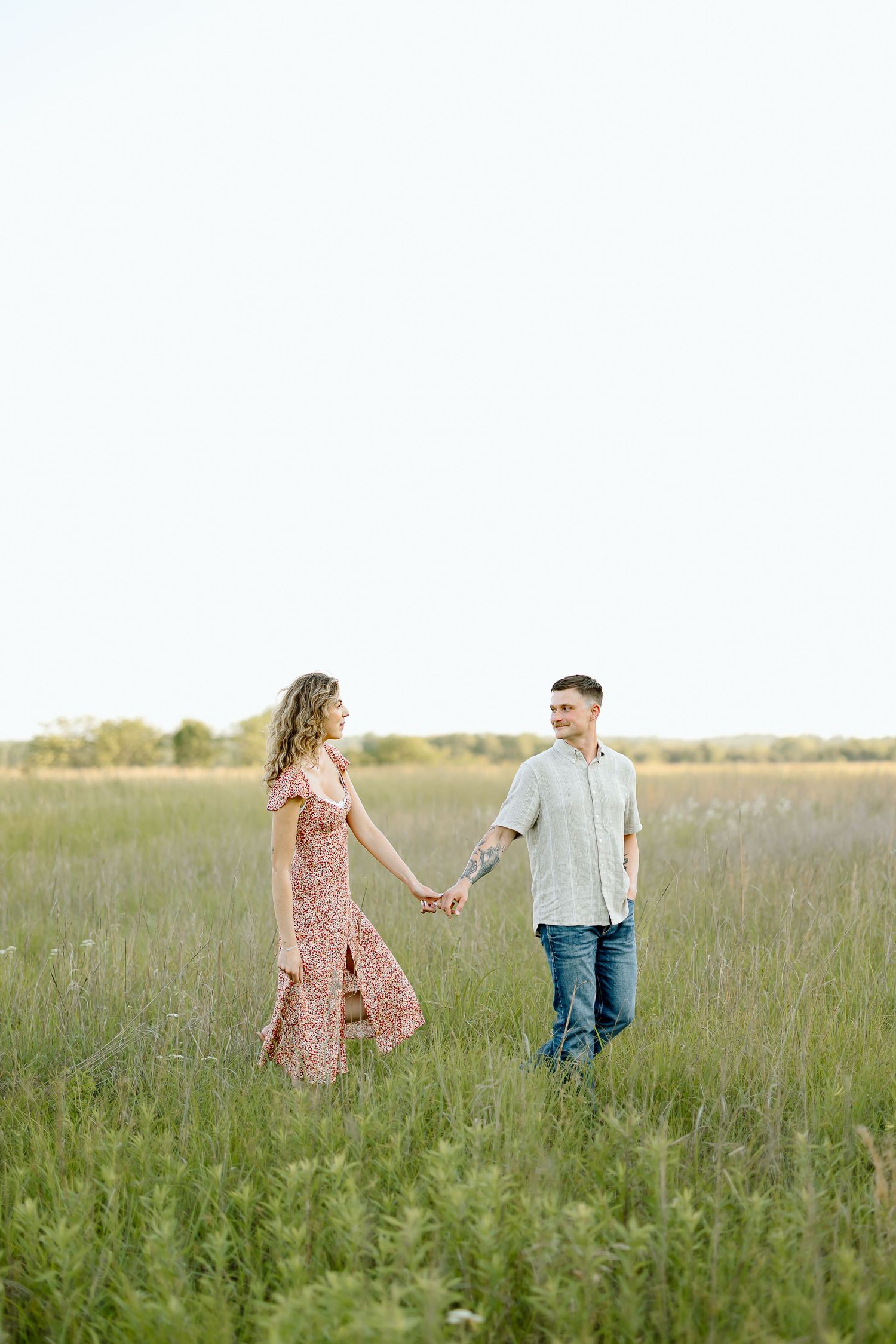 A couple walks hand in hand through a serene summer meadow wearing casual attire at sunset.