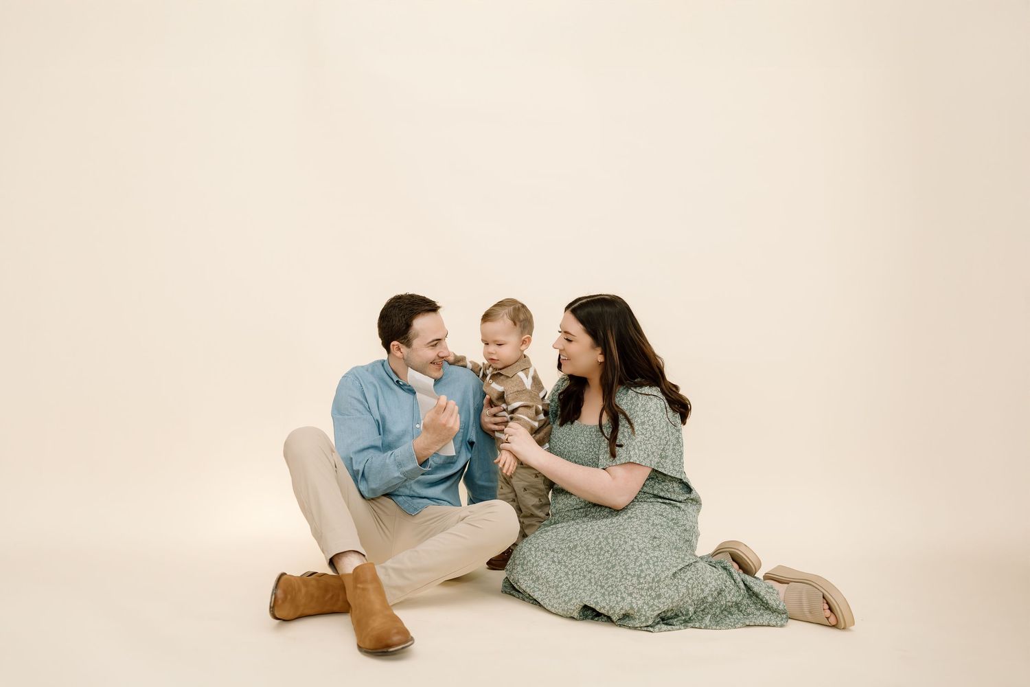 Family wearing coordinated blue and sage outfits lies together on cream colored floor in studio setting.