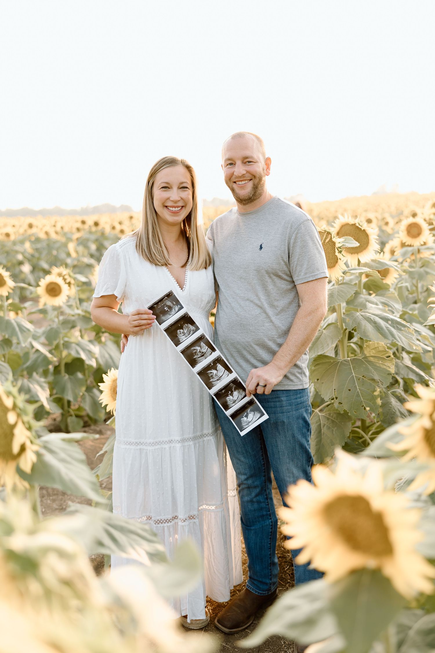A couple shares joyful moments in a sunlit sunflower field while holding ultrasound photos.