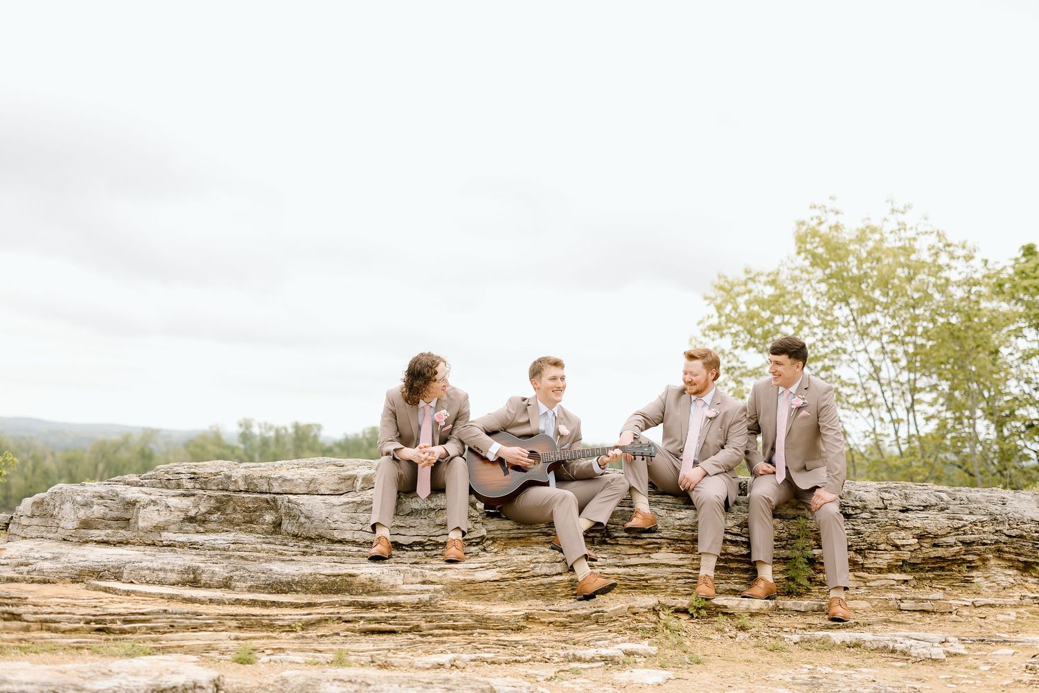 A group of people in neutral colored clothing sit casually on a rocky ledge overlooking a scenic landscape.
