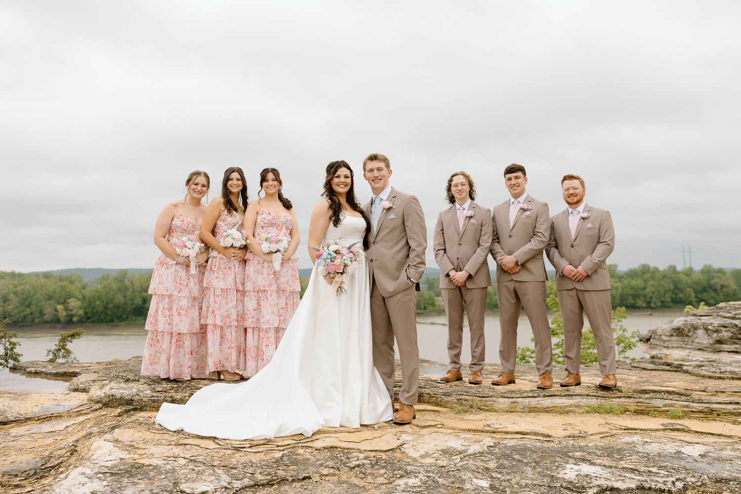Wedding party in blush pink dresses and tan suits poses together on rocky outcrop overlooking scenic landscape.