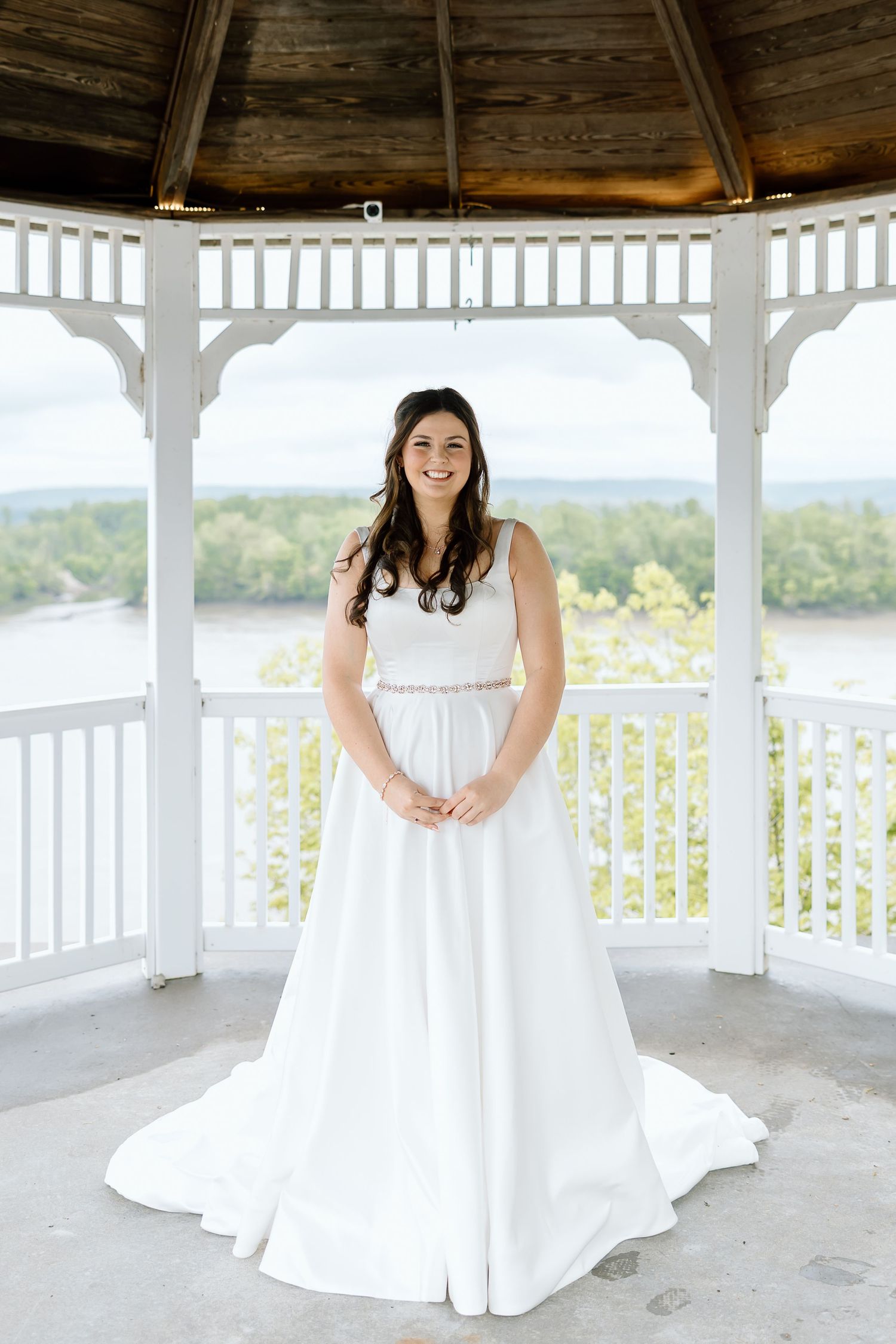 A bride in a white wedding dress poses in a scenic gazebo overlooking water and trees.