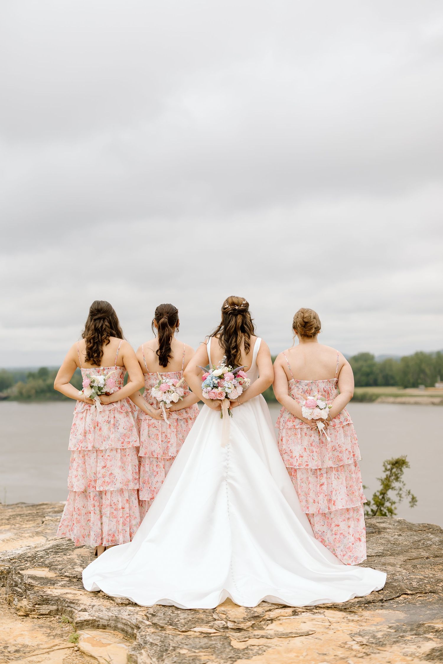 Bridal party in pink floral dresses sits with bride in white gown on rocky overlook by lake under cloudy sky.