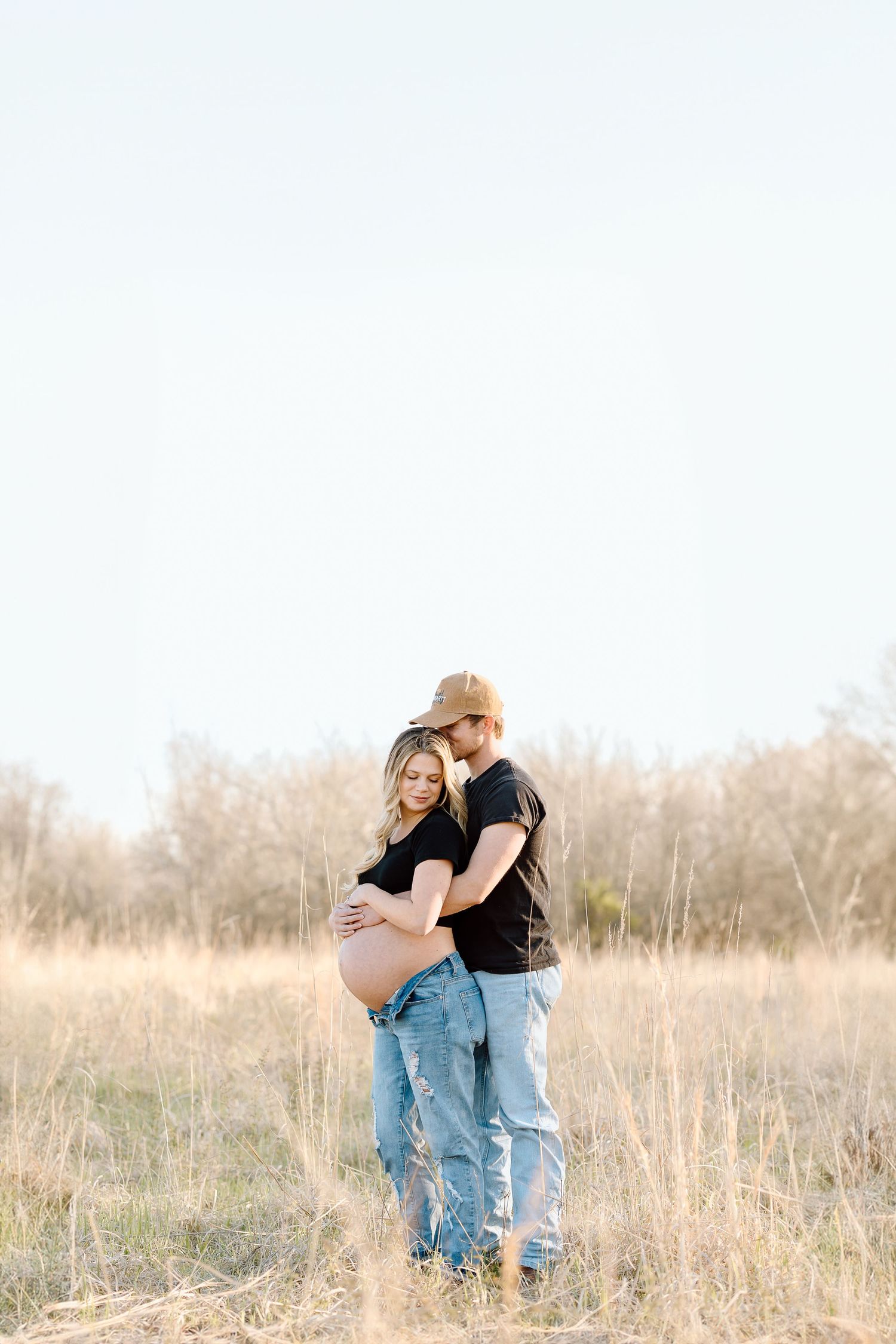 Couple embraces in a golden field during sunset with tall grass and trees in background.