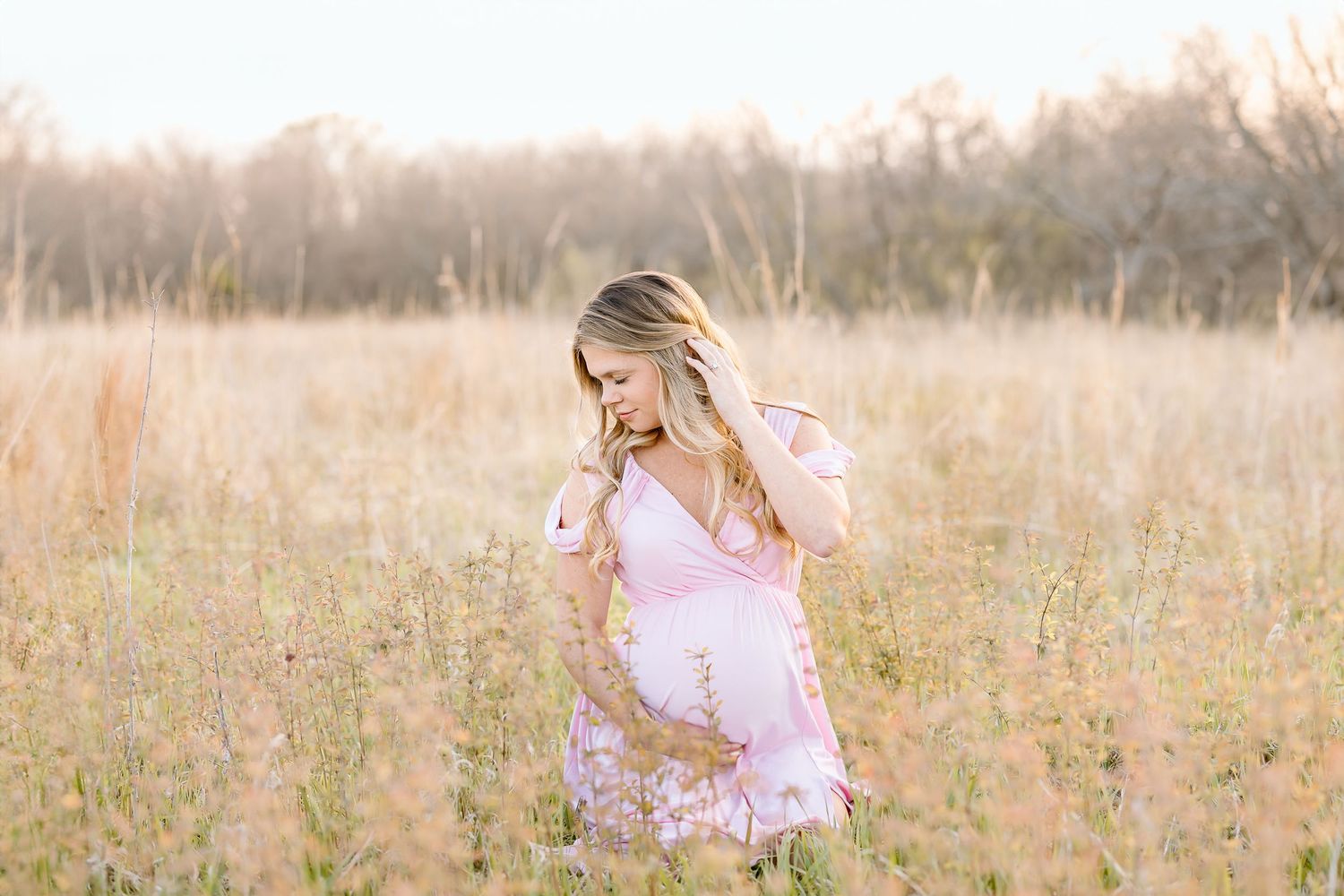 A person in a pink sweater stands in a golden autumn field during sunset with tall grass and trees in the background.