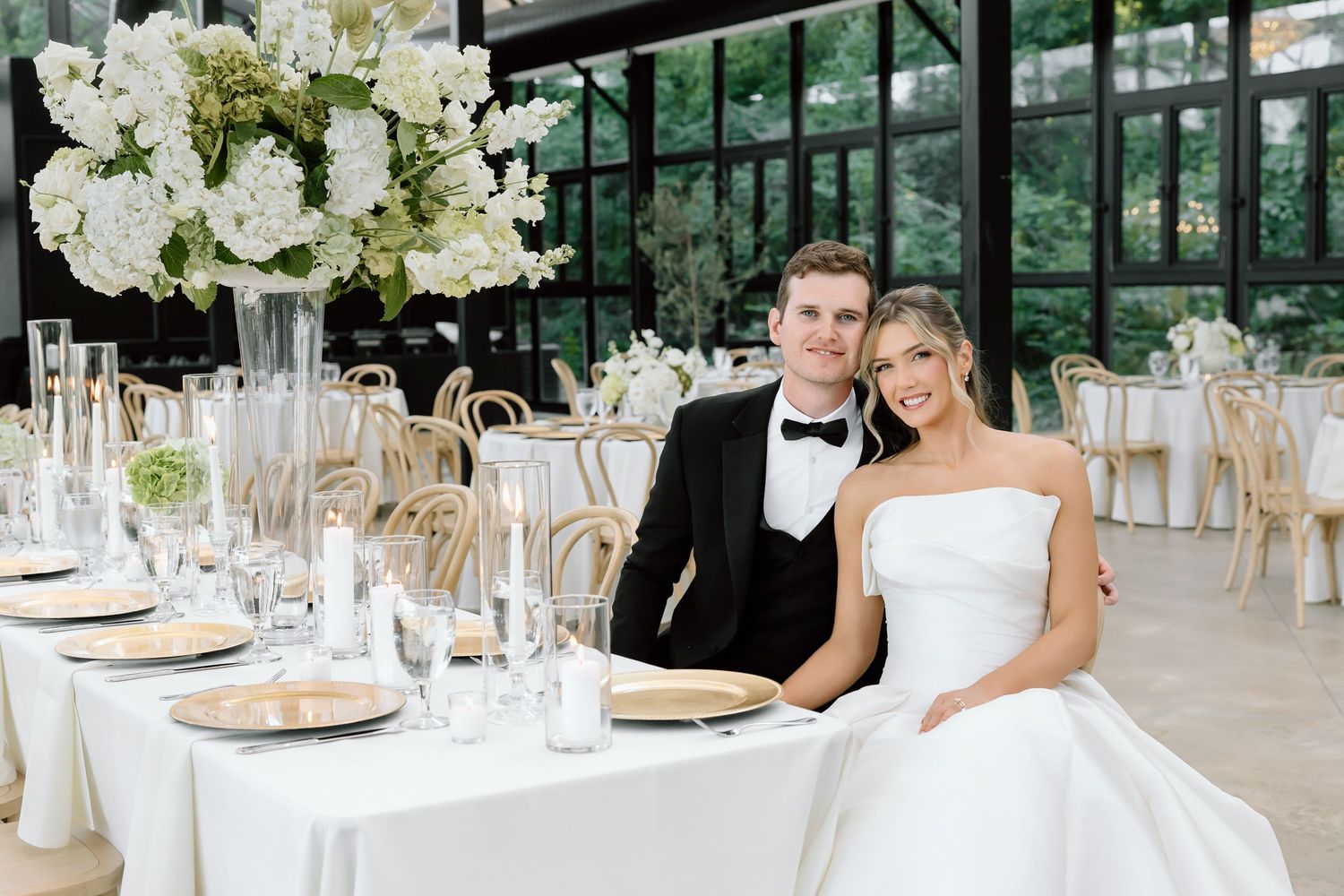 Bride and groom smiling at elegant wedding reception table with white floral centerpieces and candlelight.
