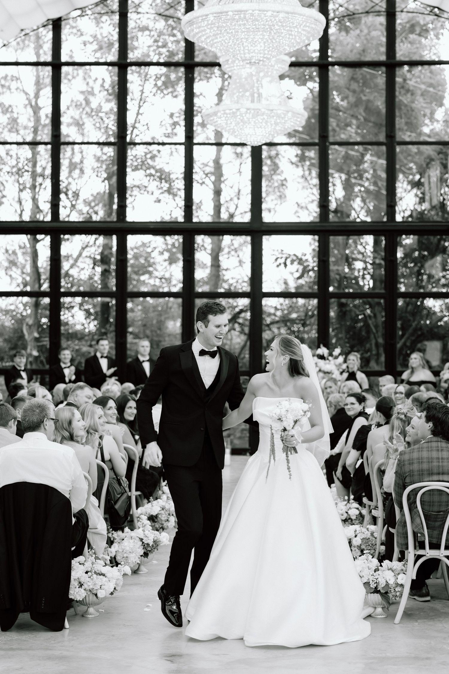 Bride and groom walk down the aisle in a stunning glass-walled venue with chandelier and floral arrangements.
