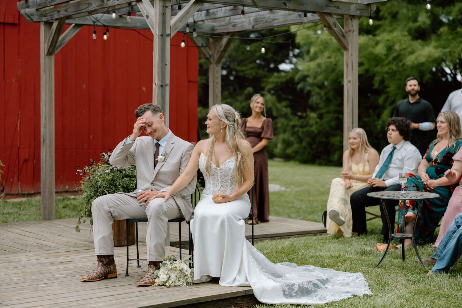 Bride and groom seated during outdoor wedding ceremony near red barn, surrounded by guests on a lush green lawn.
