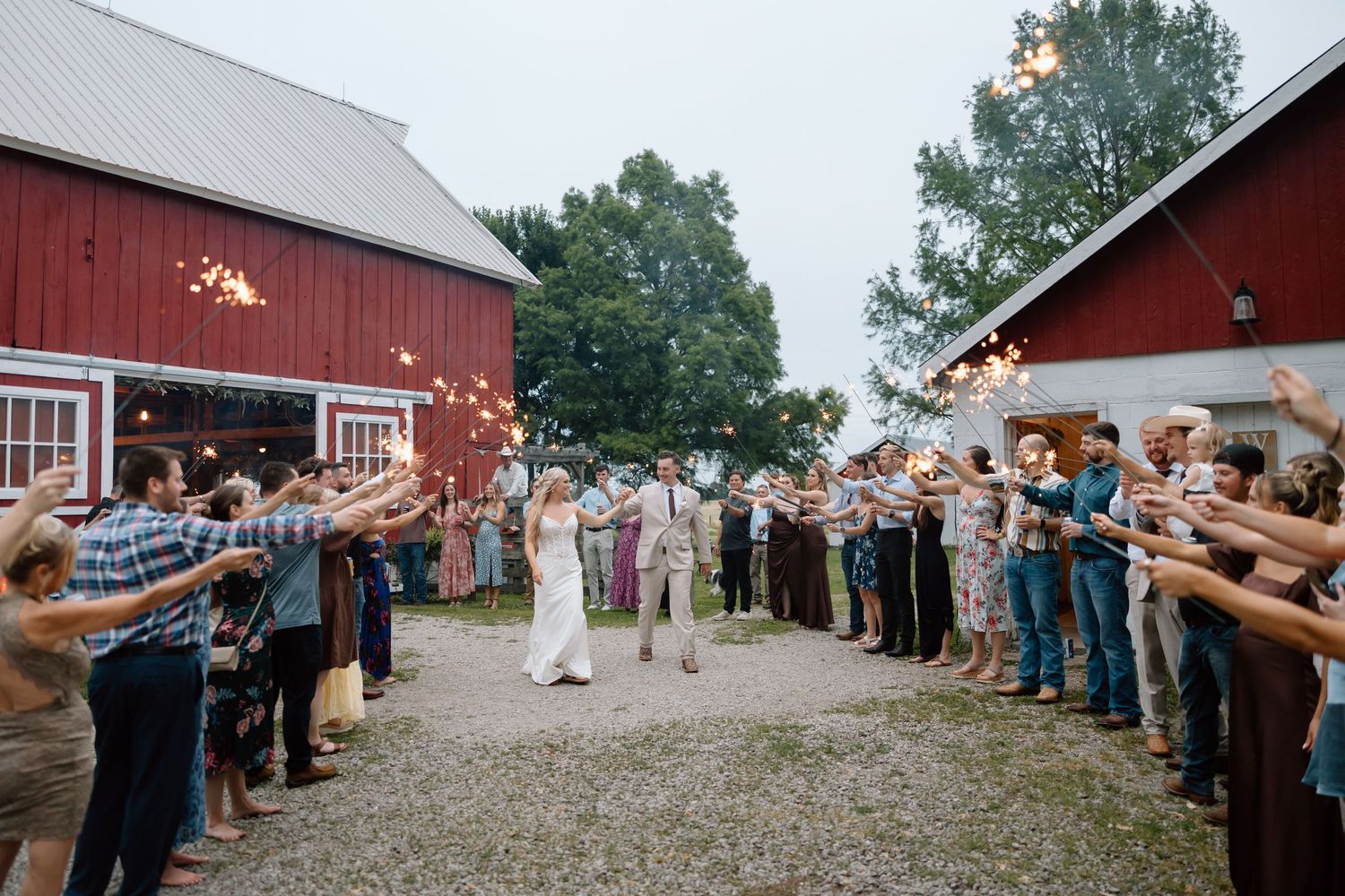 Newlywed couple exits barn wedding venue through sparkler send-off lined with cheering guests at rustic outdoor celebration.