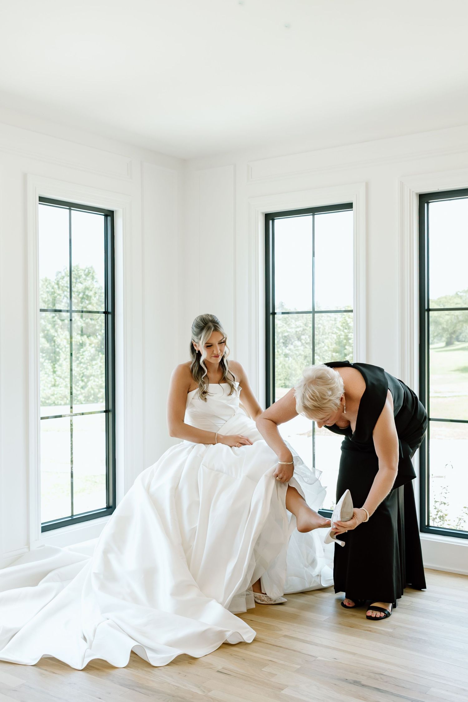 A bride in a white gown sits as a woman in black helps her put on her wedding shoes in a bright, windowed room.