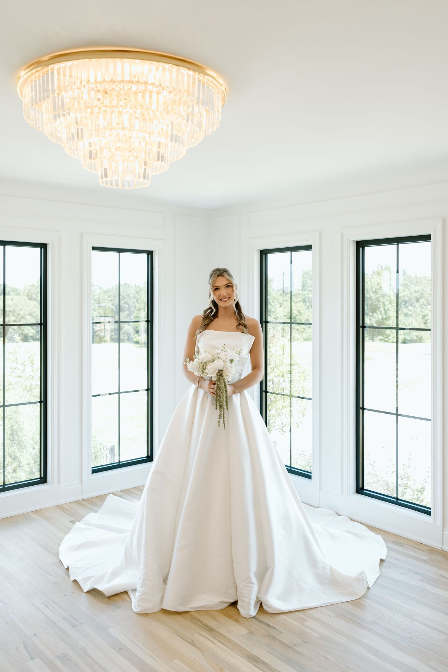 Bride in strapless white ballgown holding bouquet stands in sunlit room with chandelier and tall windows.