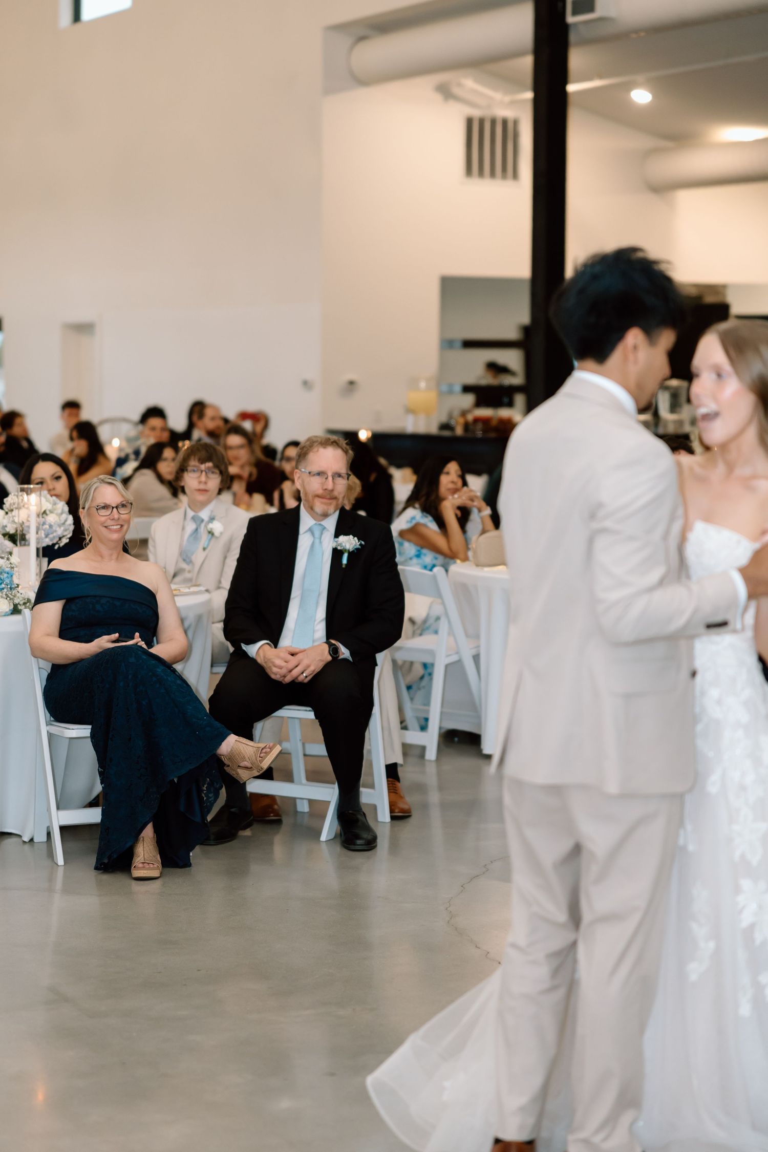 Wedding guests smile as a bride in a white gown and groom in a light suit walk past during their indoor ceremony.