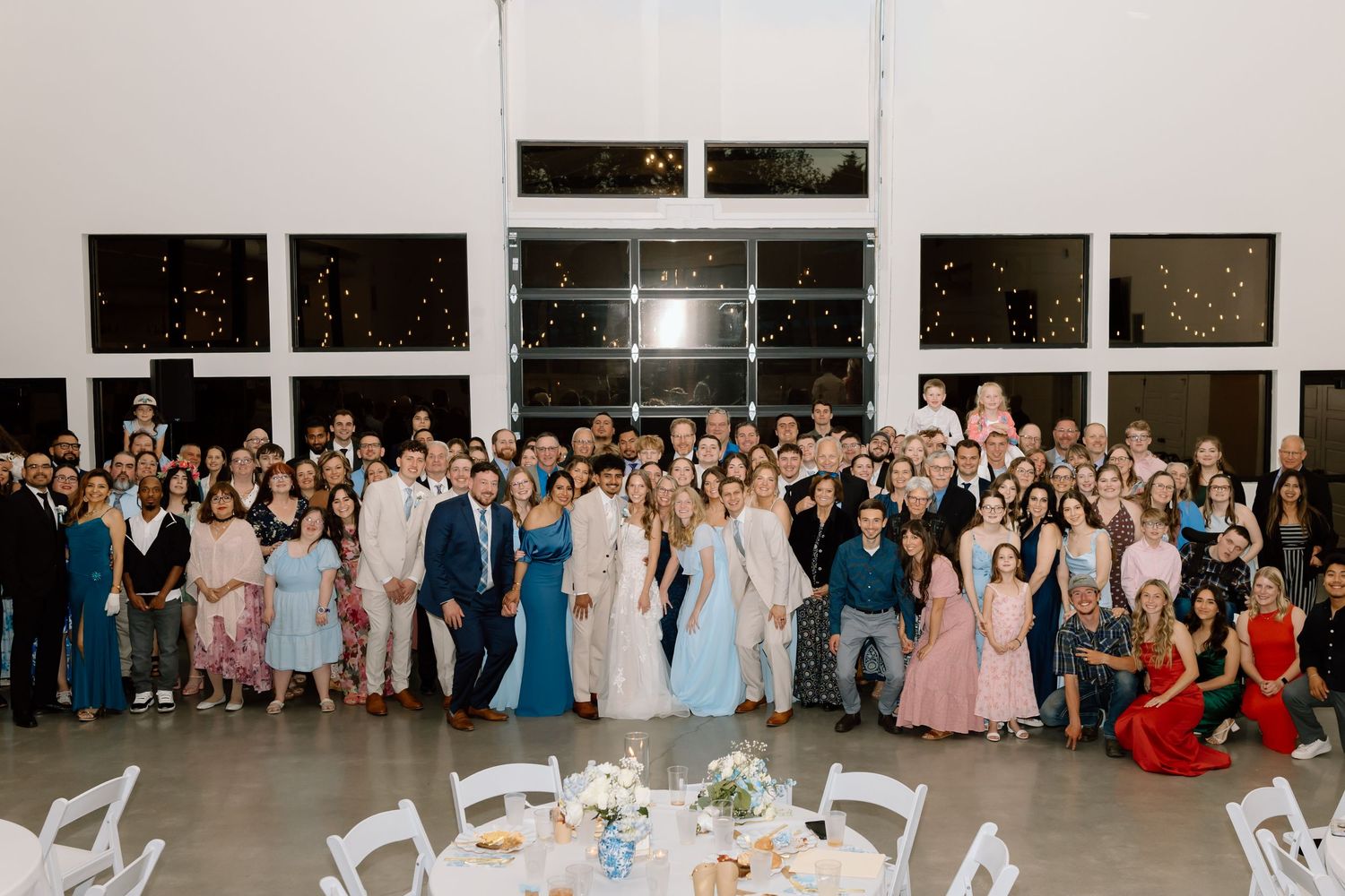 Large wedding reception group photo with guests in formal attire gathered in a modern venue with floor-to-ceiling windows.