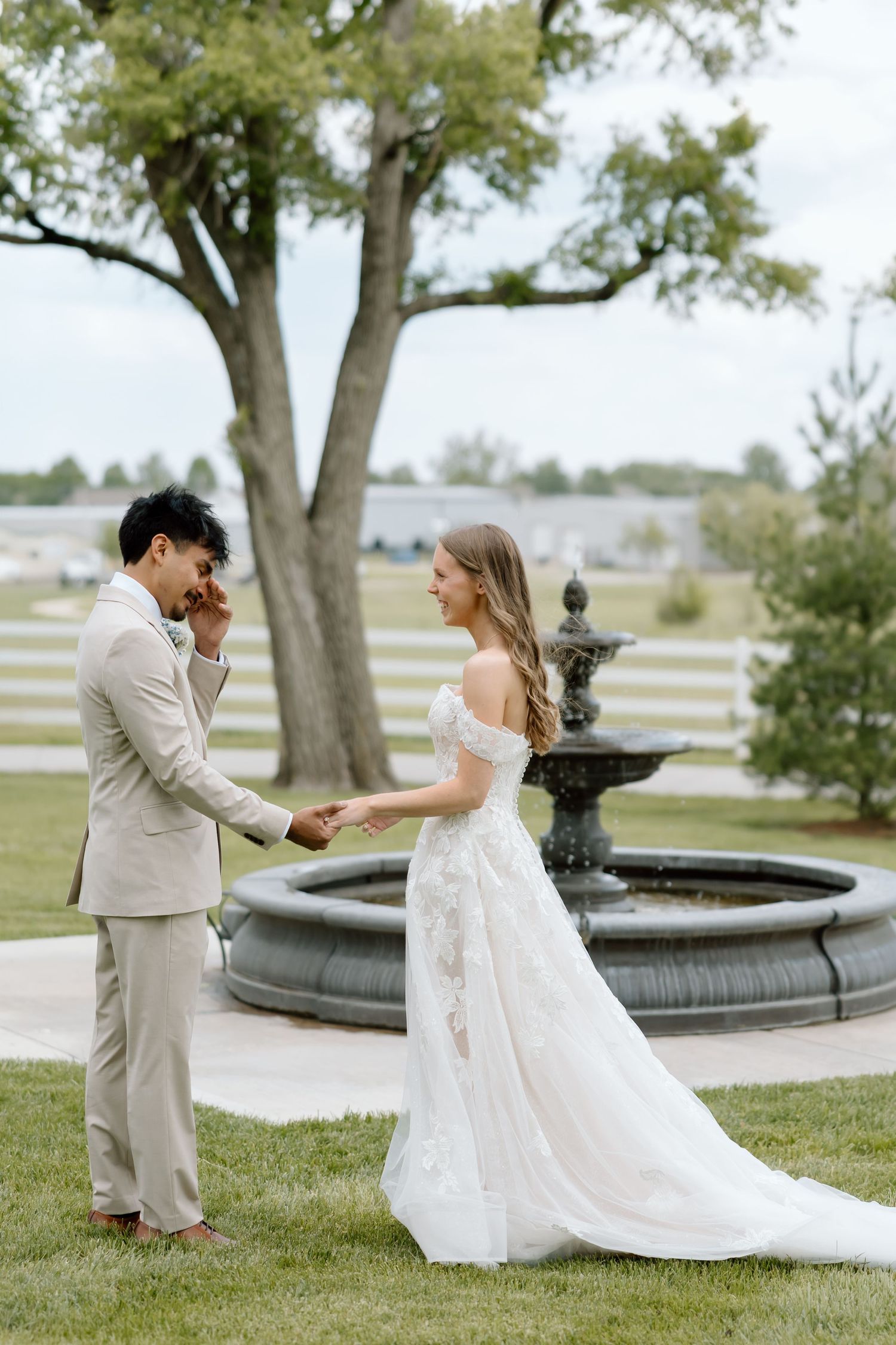 A bride and groom hold hands near a stone fountain during an outdoor wedding ceremony on a lush green lawn.