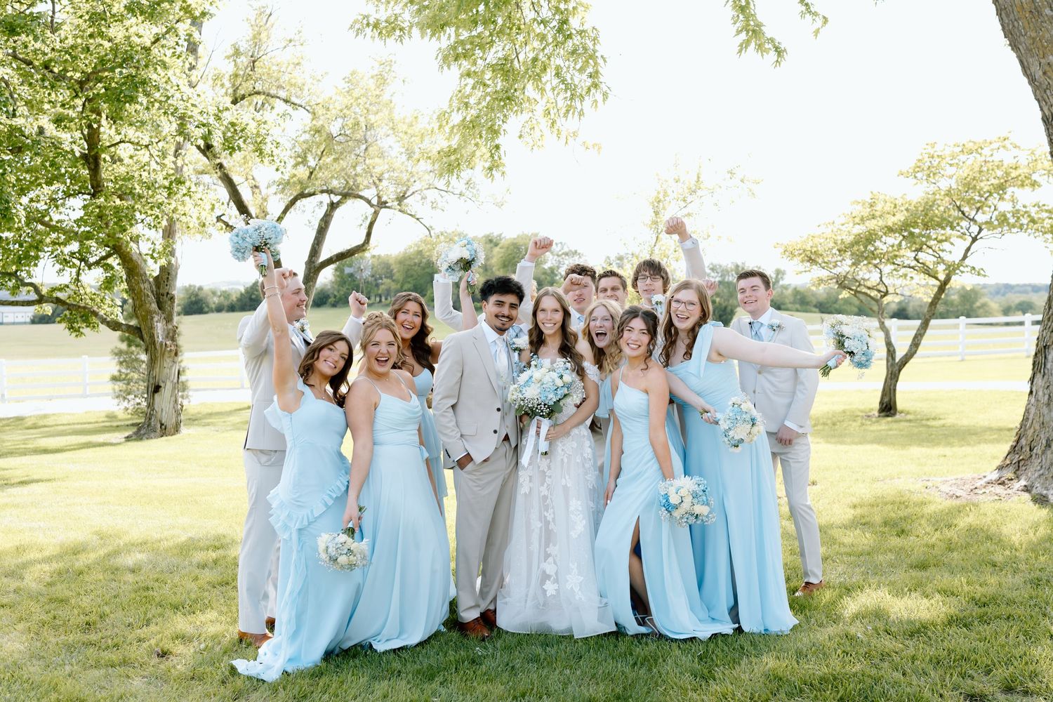 Wedding party in light blue dresses and tan suits celebrating outdoors near a lake on a sunny day.