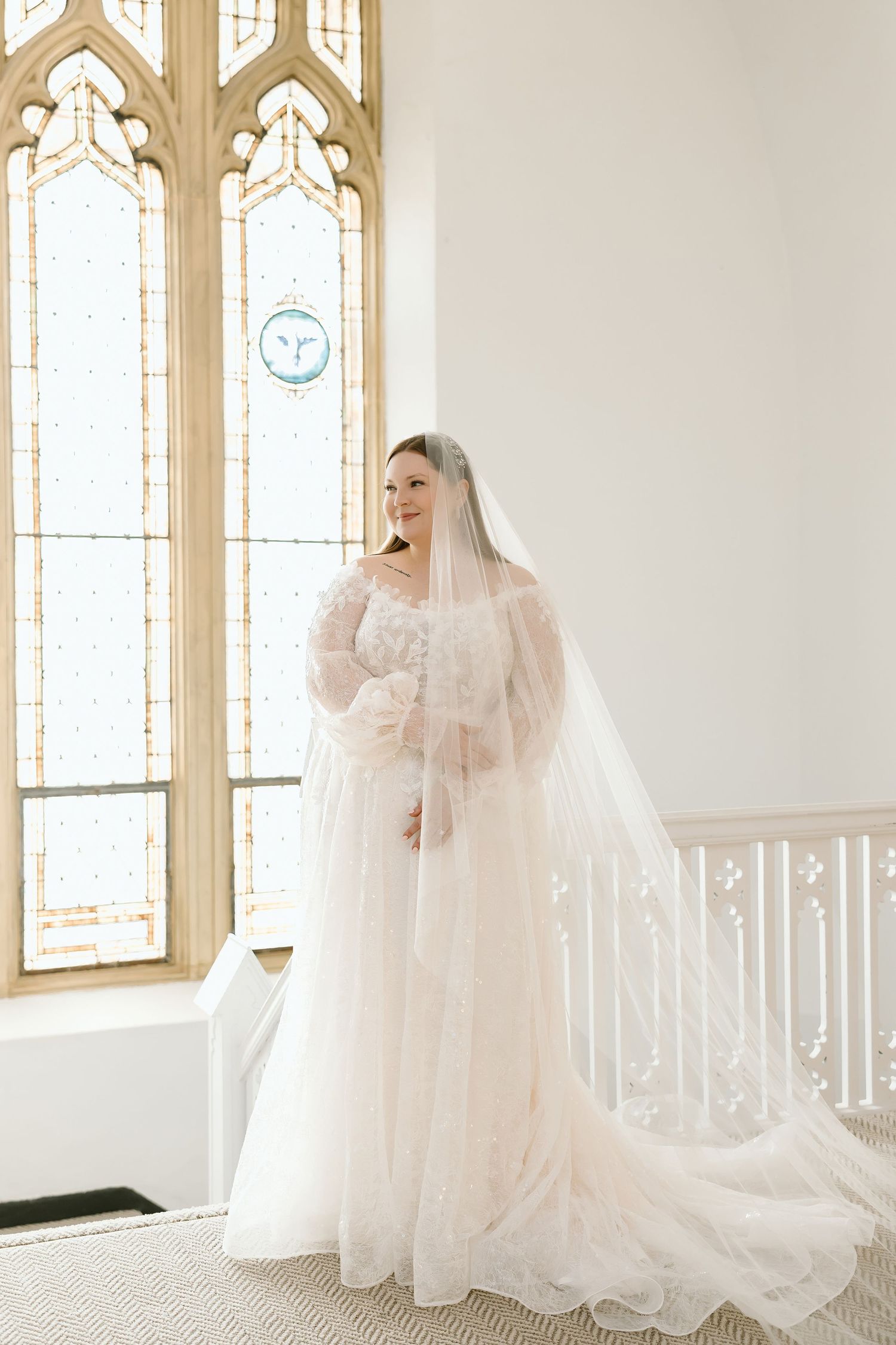 Bride in lace wedding gown and cathedral veil stands by stained glass window in elegant white chapel.