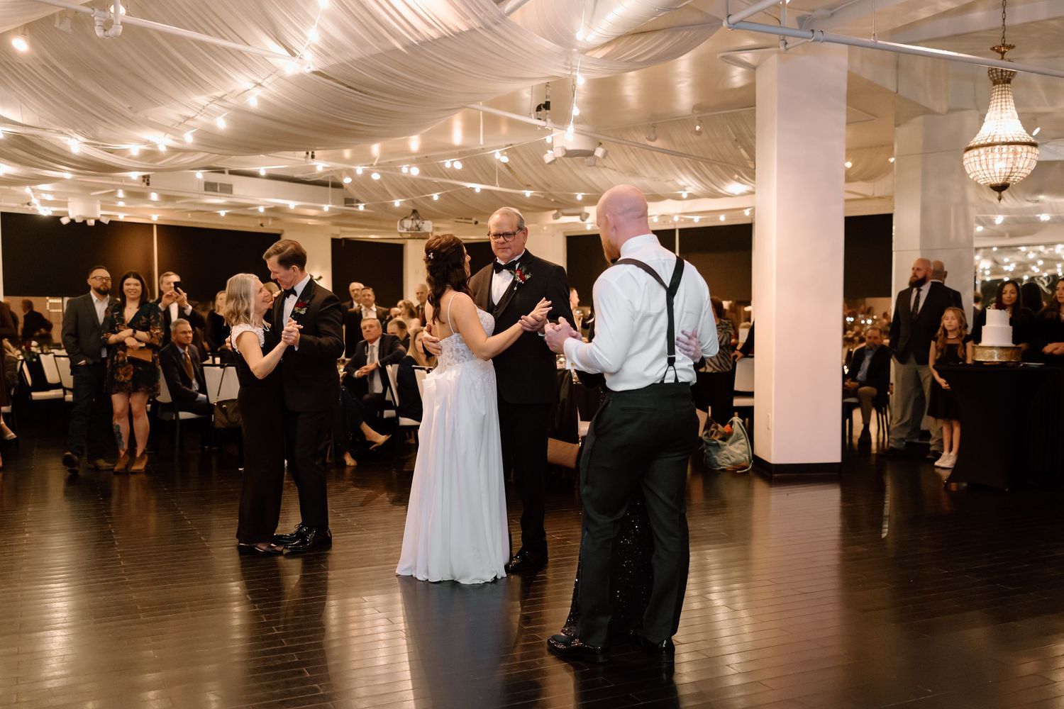 Wedding guests dance together in an elegant ballroom reception venue with warm lighting and white decor.