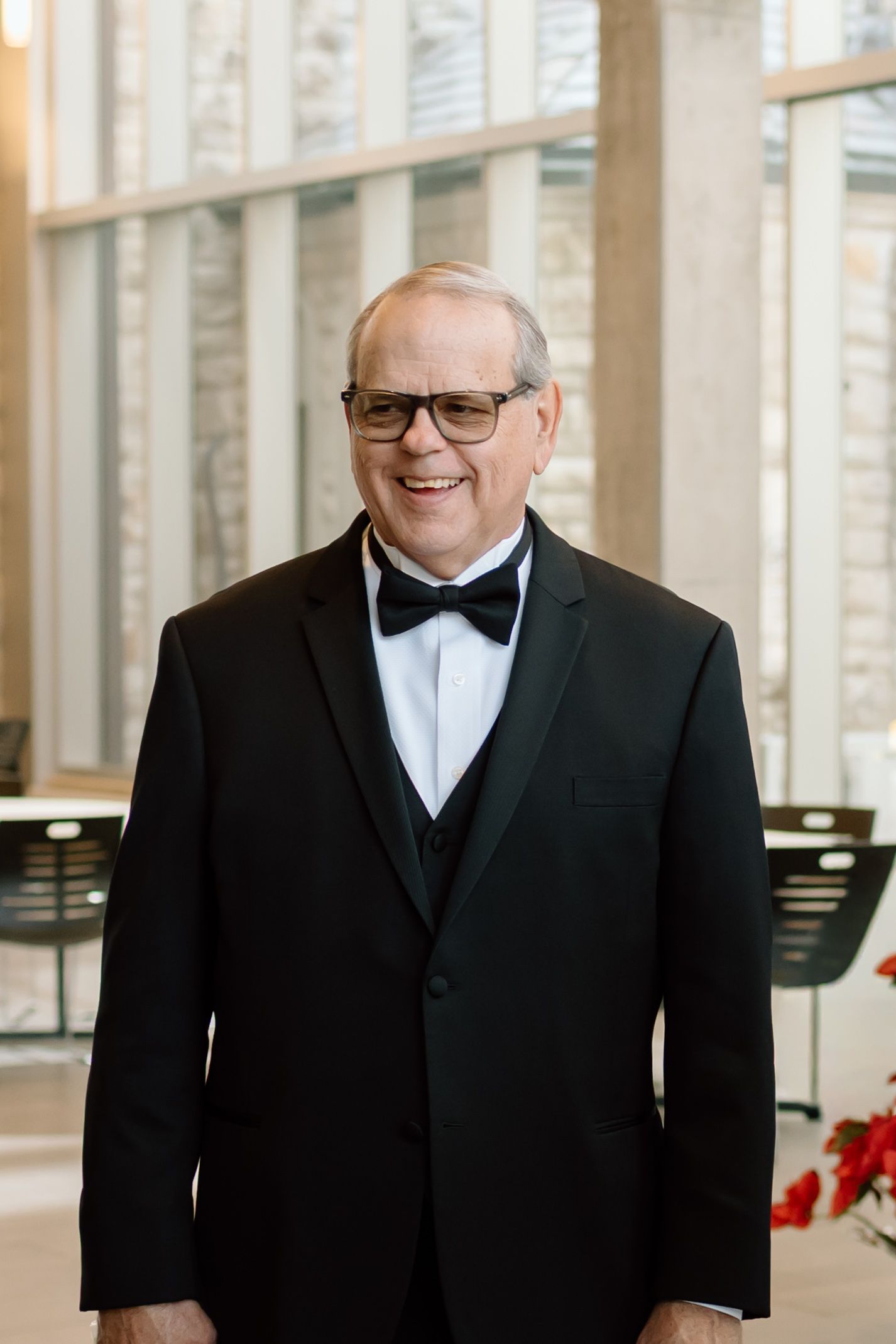 A person wearing a black tuxedo and bow tie smiles warmly in an elegant indoor setting.
