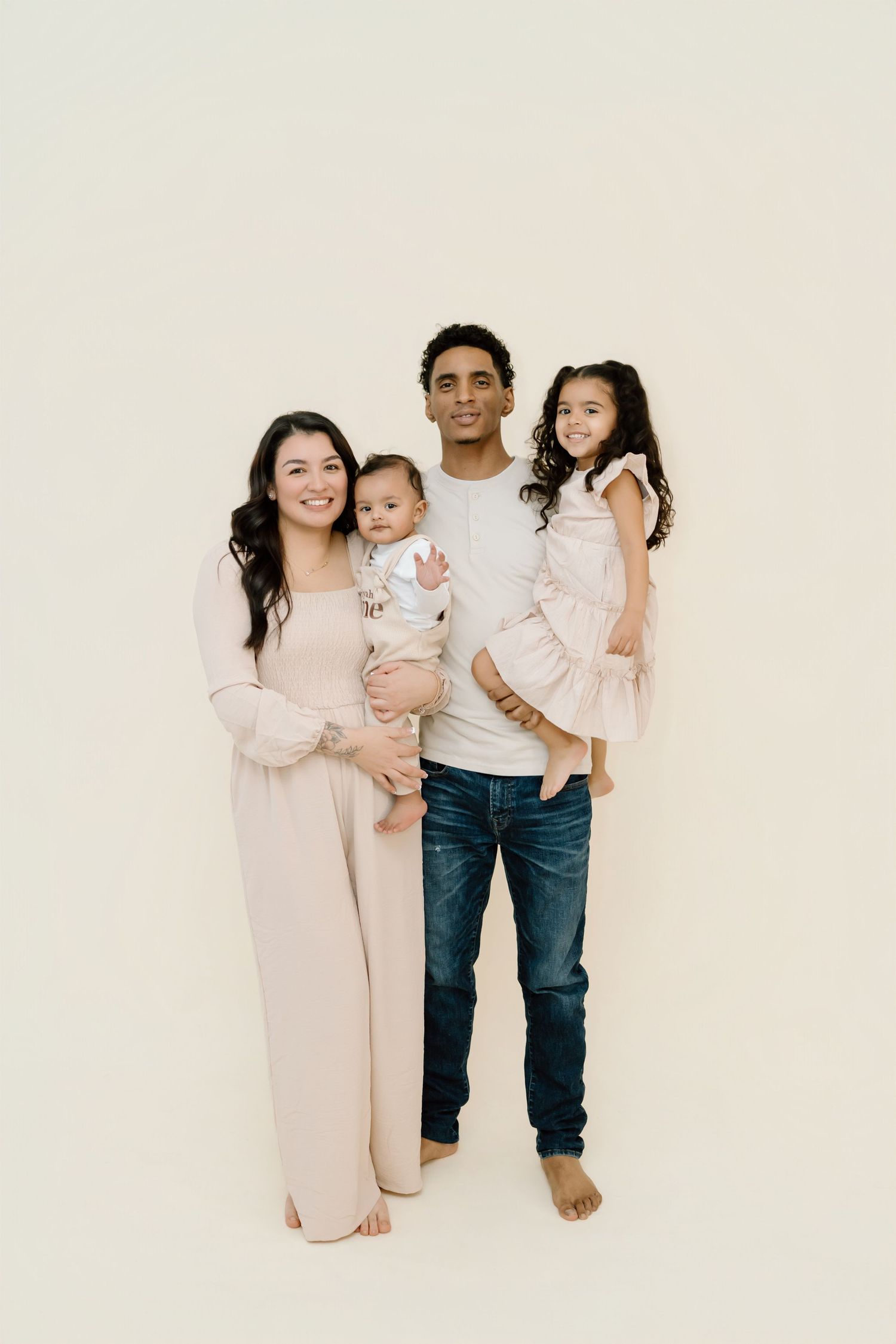 A family wearing light pink and white outfits poses together against a cream colored backdrop for a studio portrait.