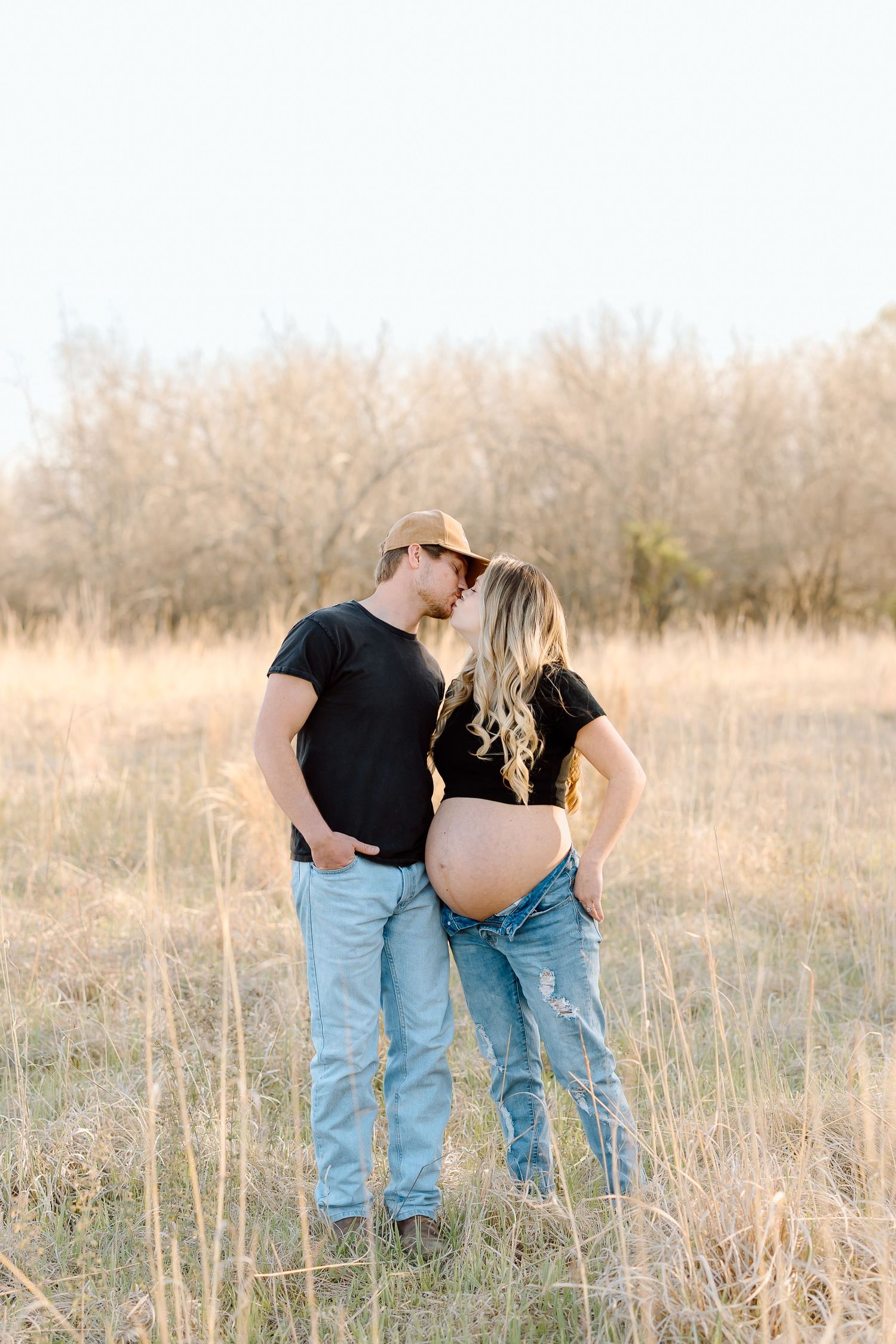 A couple in matching black shirts and blue jeans embrace while standing in a golden sunlit field at sunset.