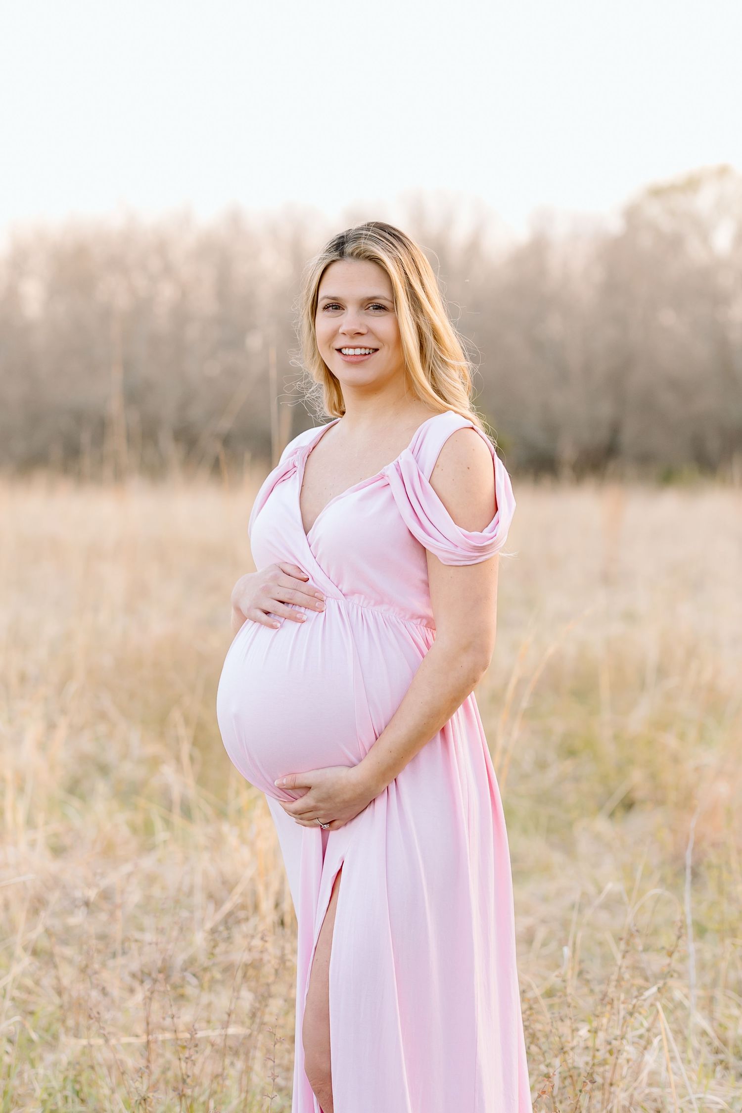 Pregnant woman in soft pink dress stands in sunlit field of tall grass at golden hour.